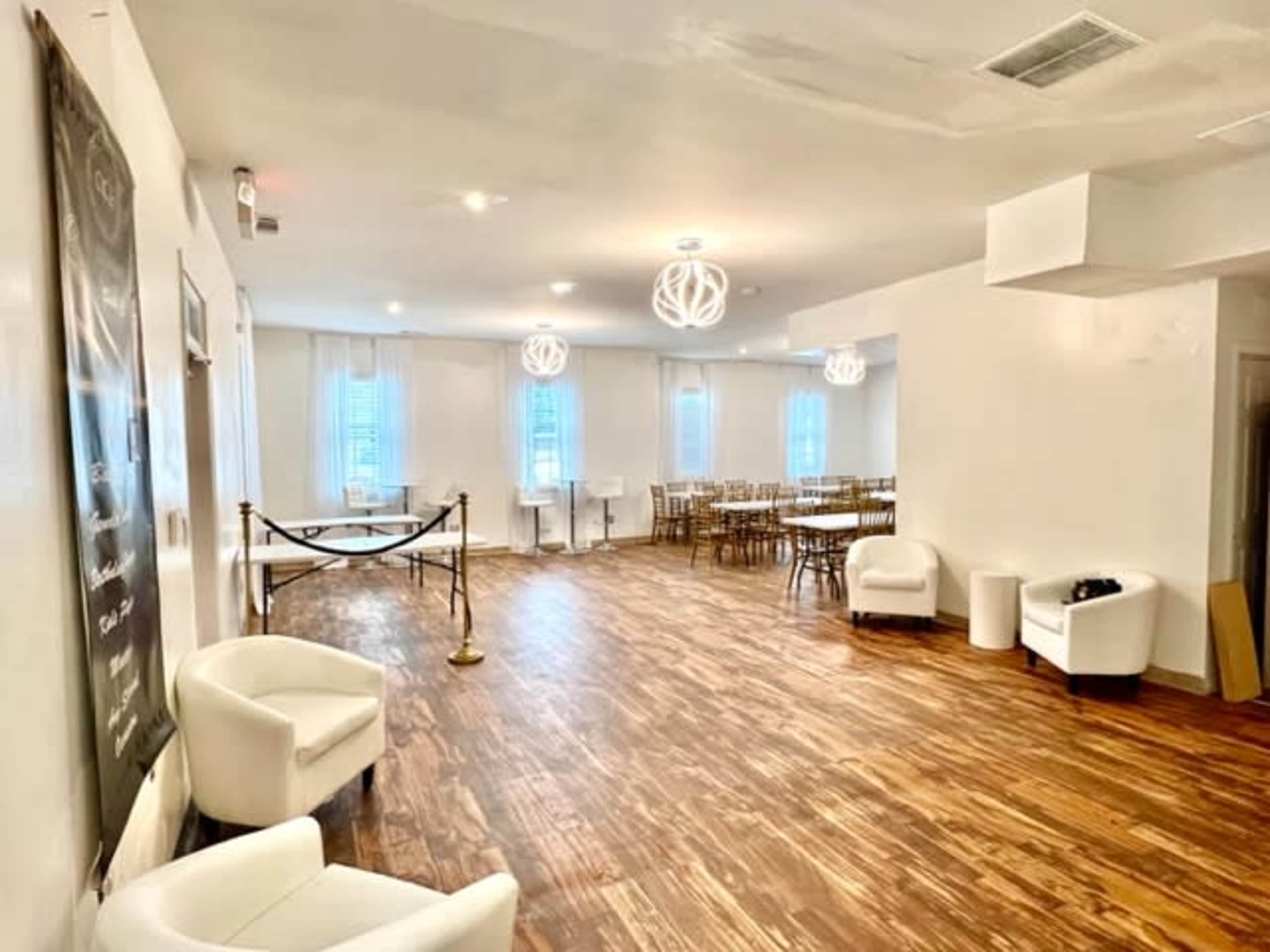 The interior of a spacious, well-lit venue features wooden flooring, white walls, and a setup of tables and chairs in a dining area.