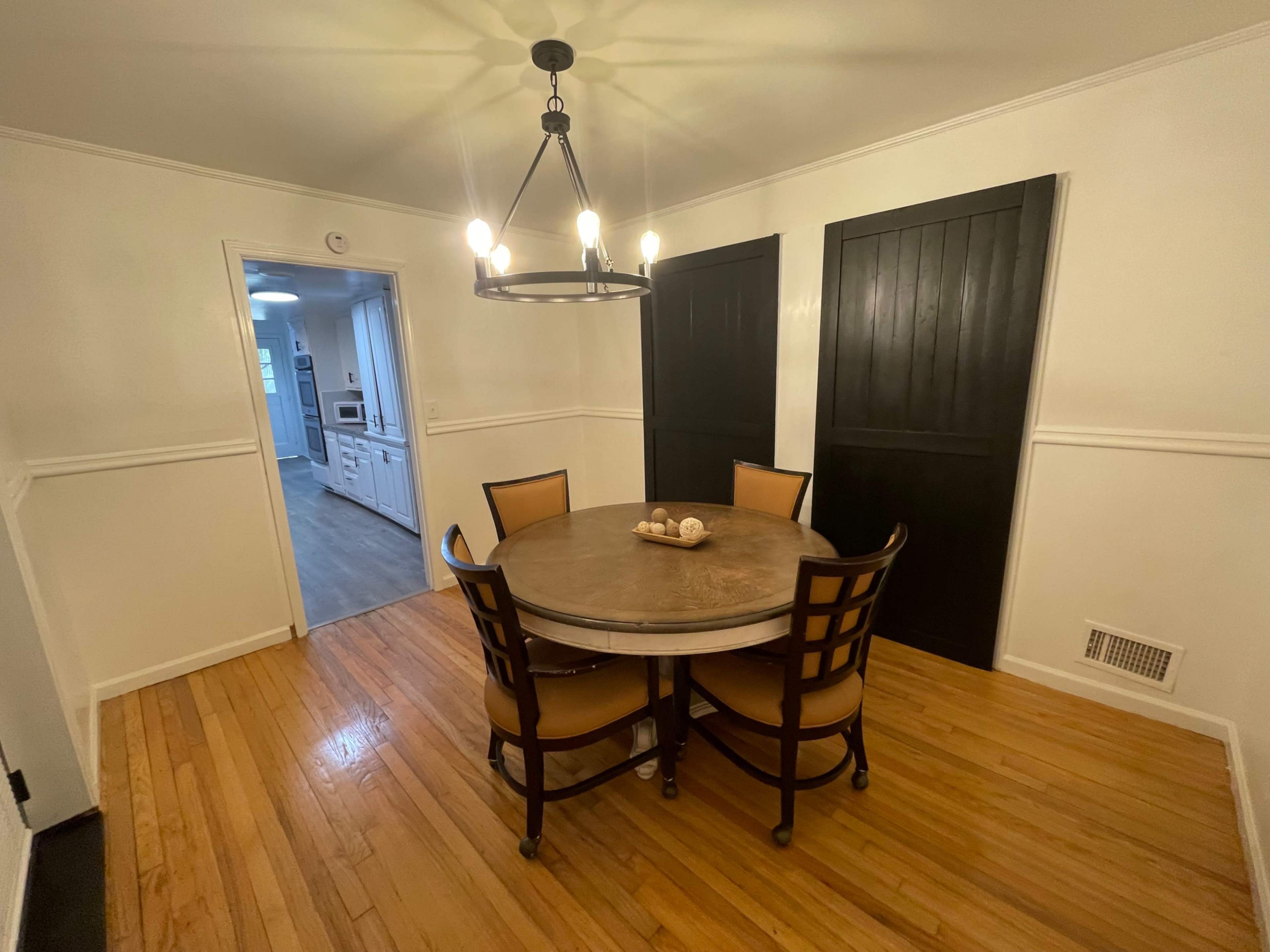 A round wooden dining table with four chairs is set in a well-lit room that features dark sliding doors and a doorway leading to a kitchen.