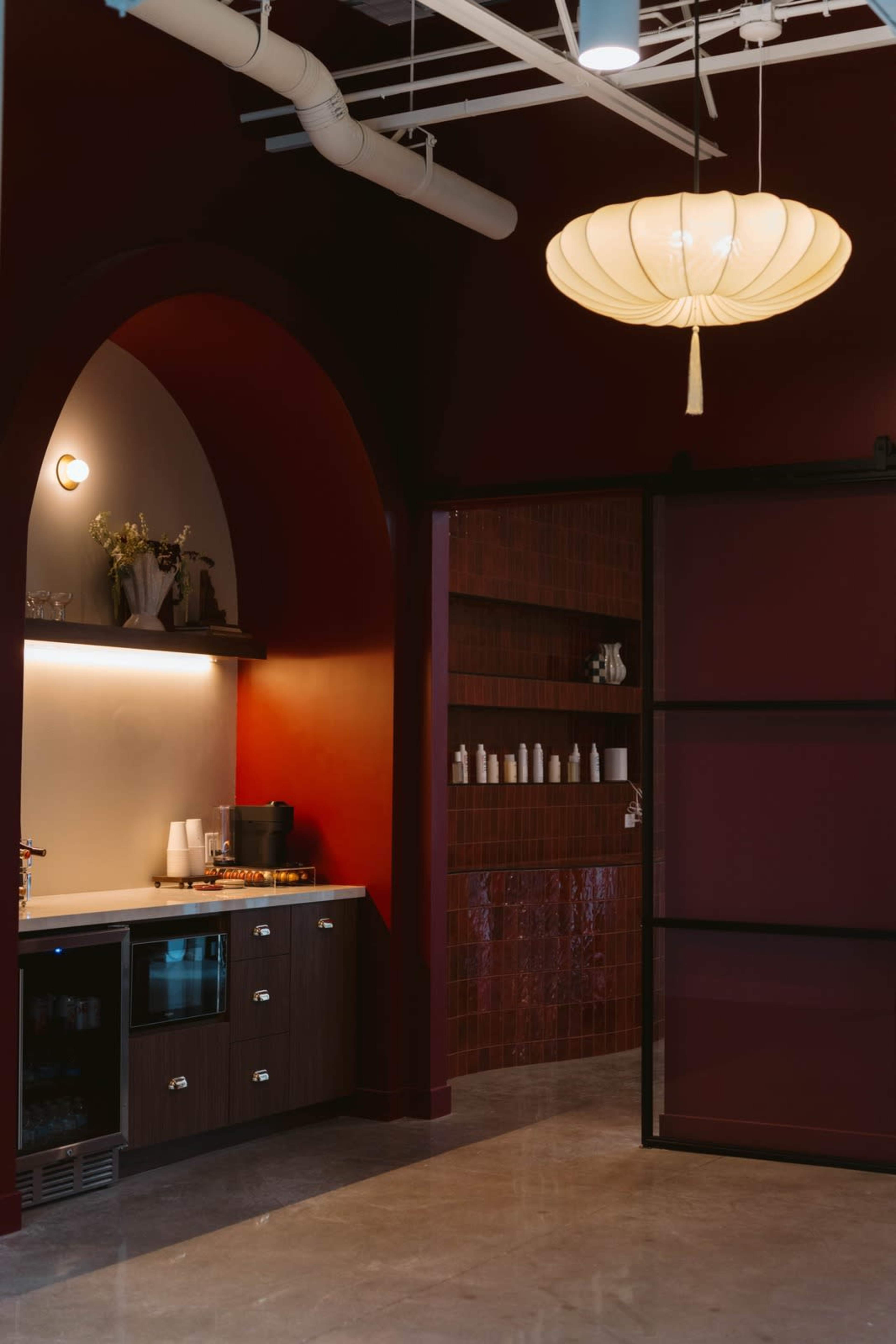 The image features a modern kitchen area with dark red walls, a countertop with an under-counter refrigerator and a few decorative items, alongside a partition leading to another section of the space.