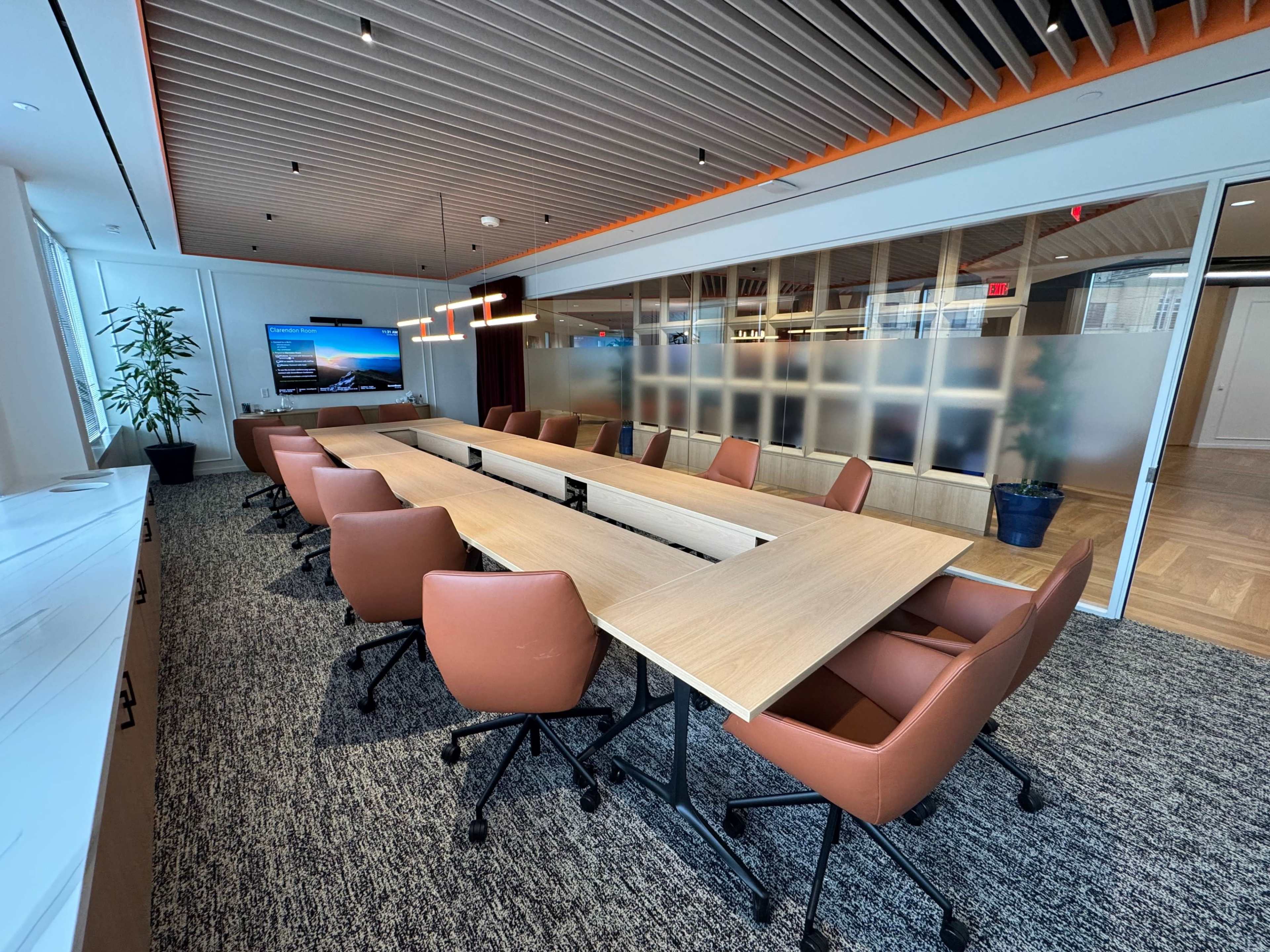 A modern conference room features a long table with eight brown chairs, large windows providing natural light, and a display screen on the wall.