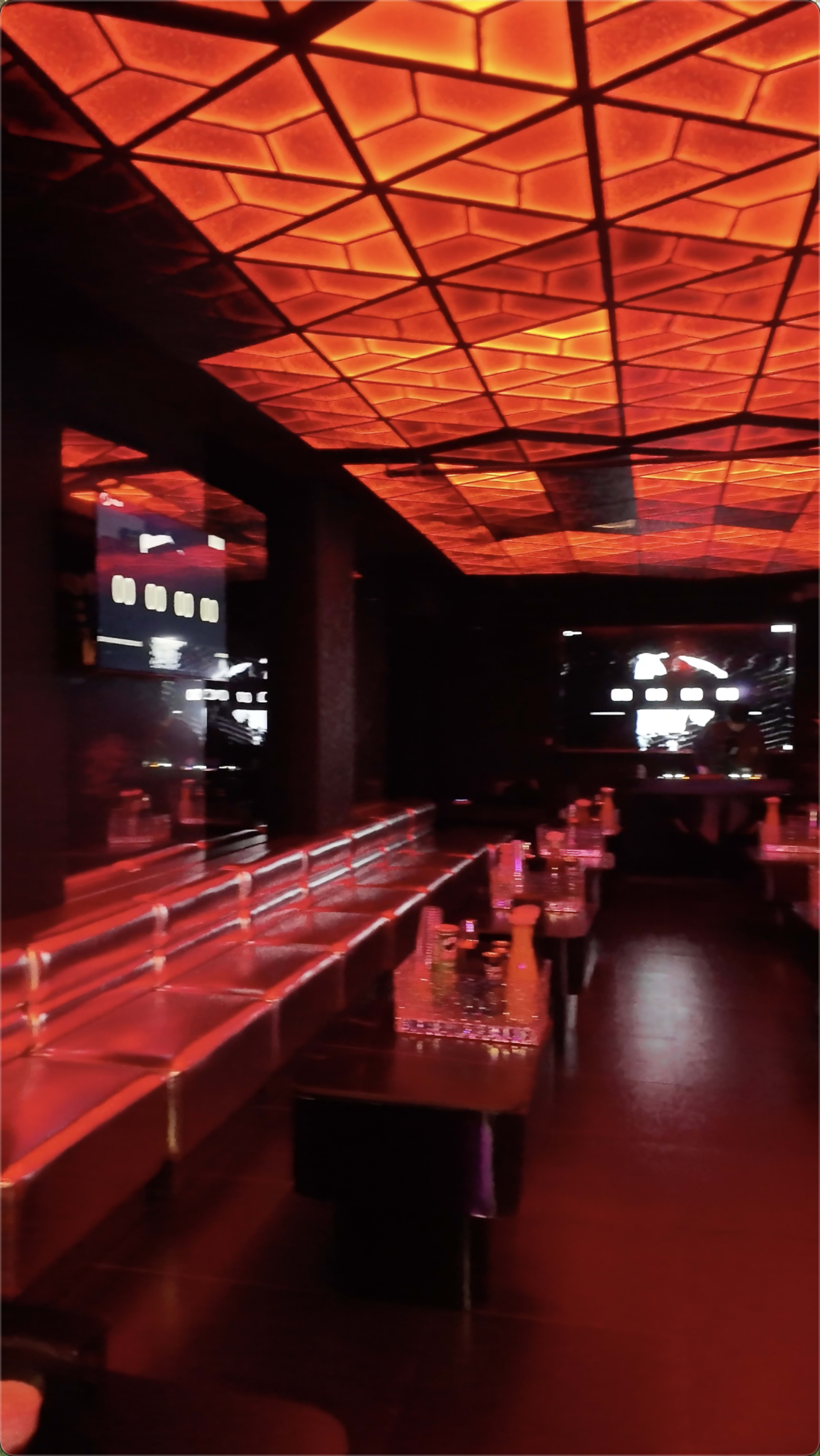 The image shows a dimly lit lounge or nightclub with a geometric, illuminated ceiling and sleek seating areas.
