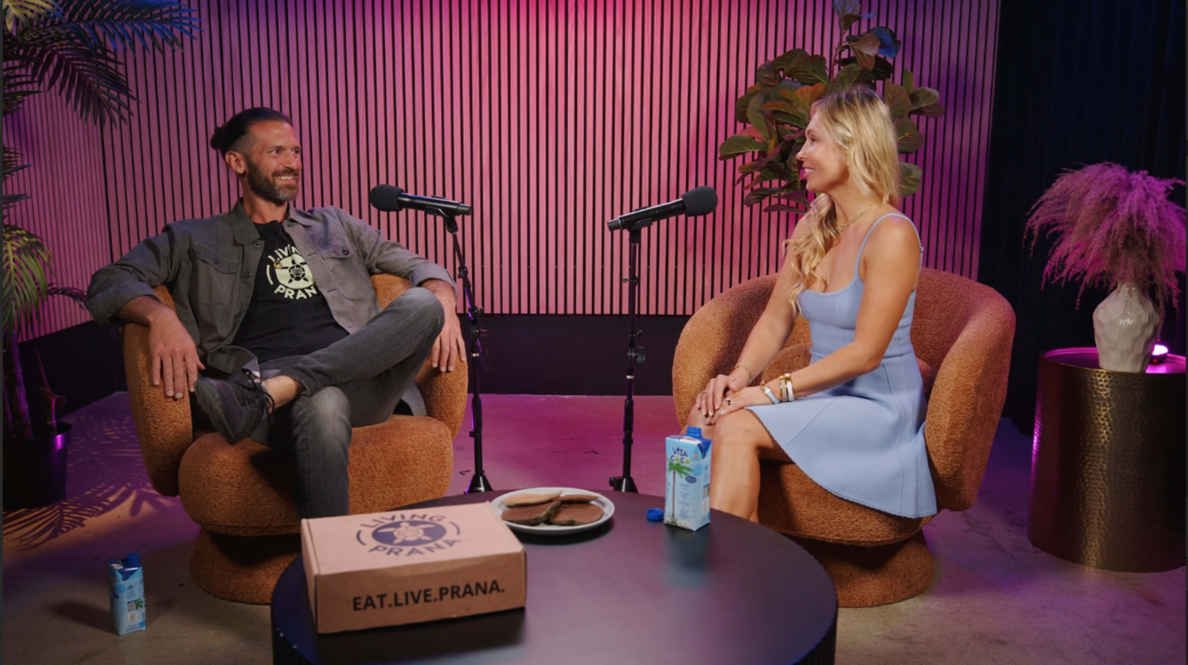 Two individuals engage in conversation while seated in modern, curved chairs next to a small round table with snacks and drinks, under vibrant lighting.