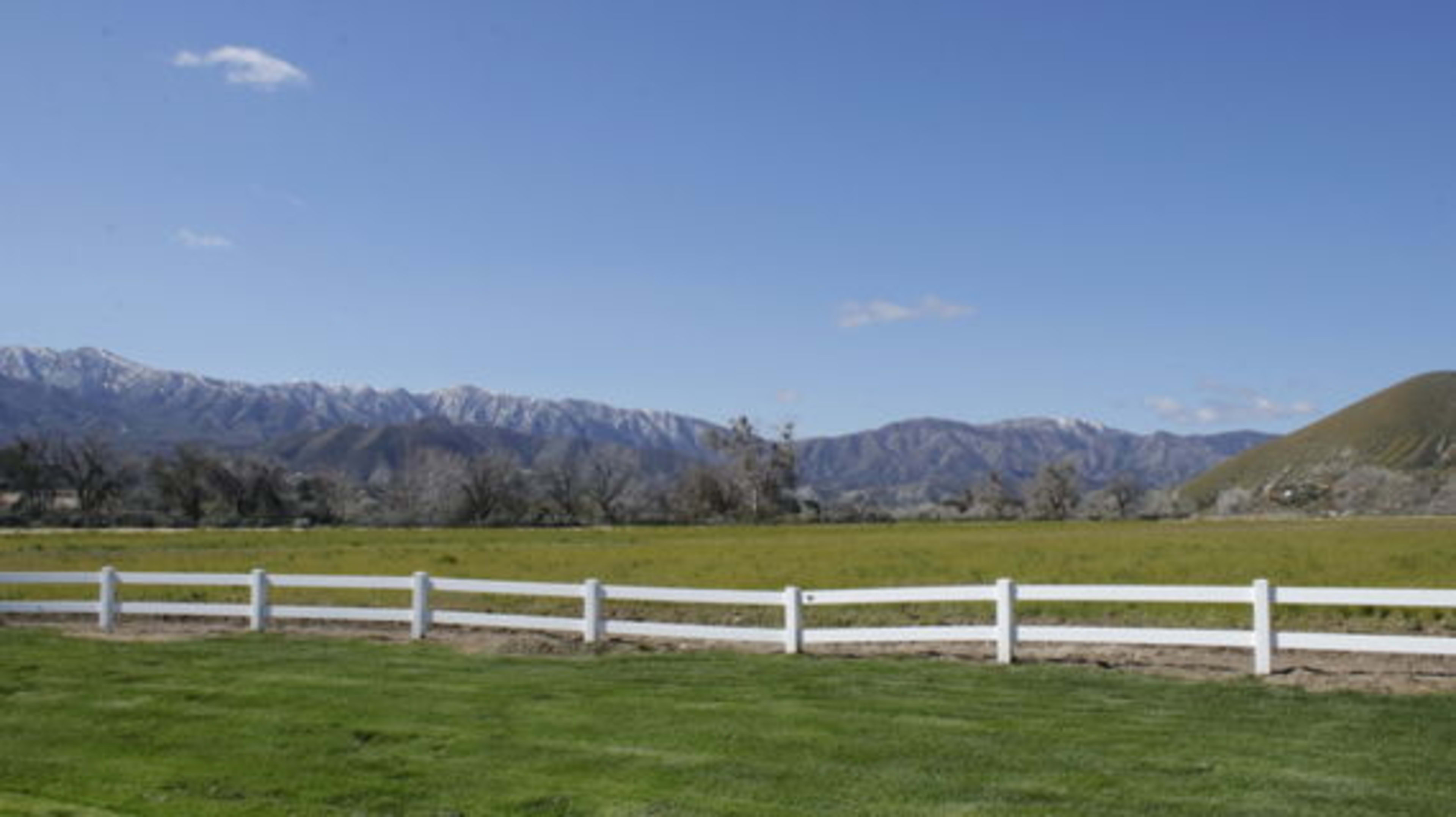 A grassy landscape with a white fence in the foreground and snow-capped mountains in the distance under a clear blue sky.