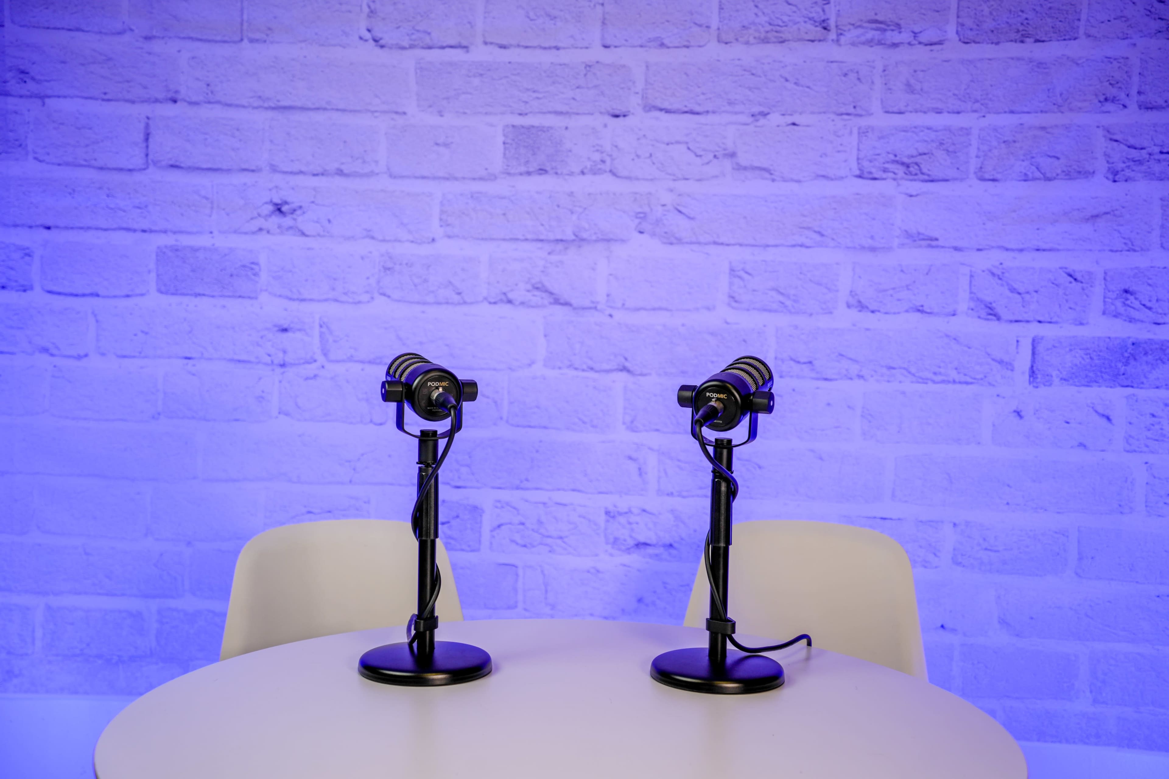 Two microphones are positioned on stands atop a round table in front of a blue-lit brick wall.