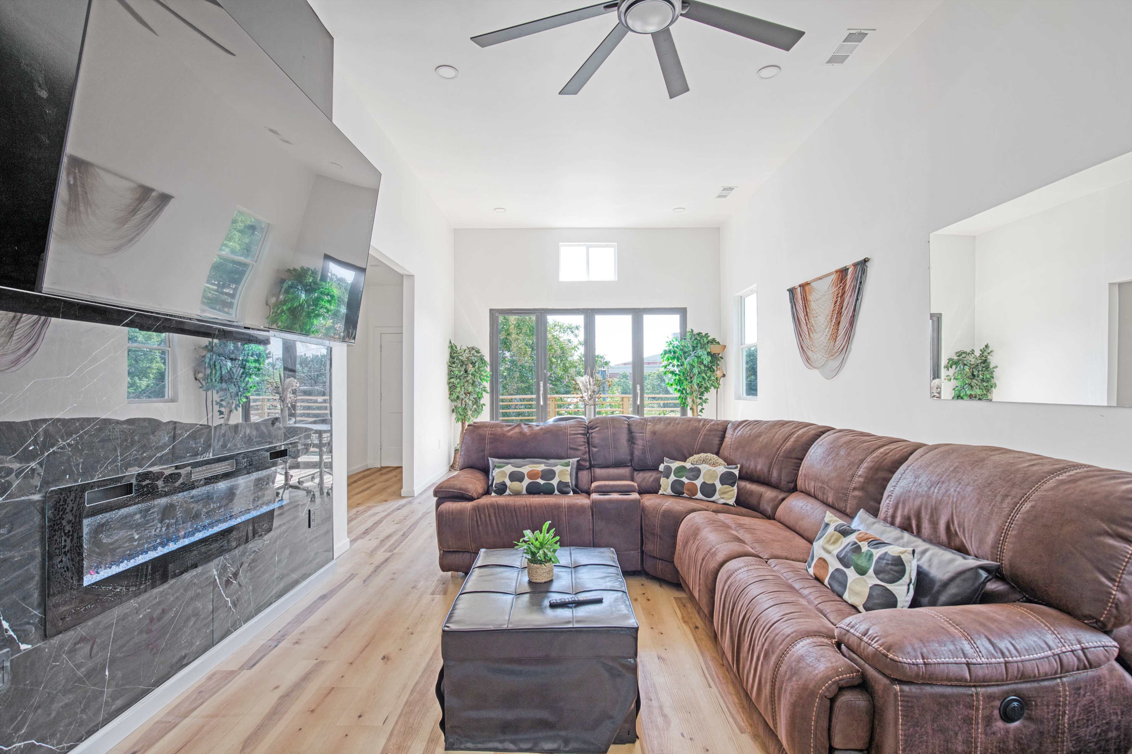 The image shows a modern living room with a brown sectional couch, a mounted flat-screen TV, a faux fireplace, and large windows allowing natural light.