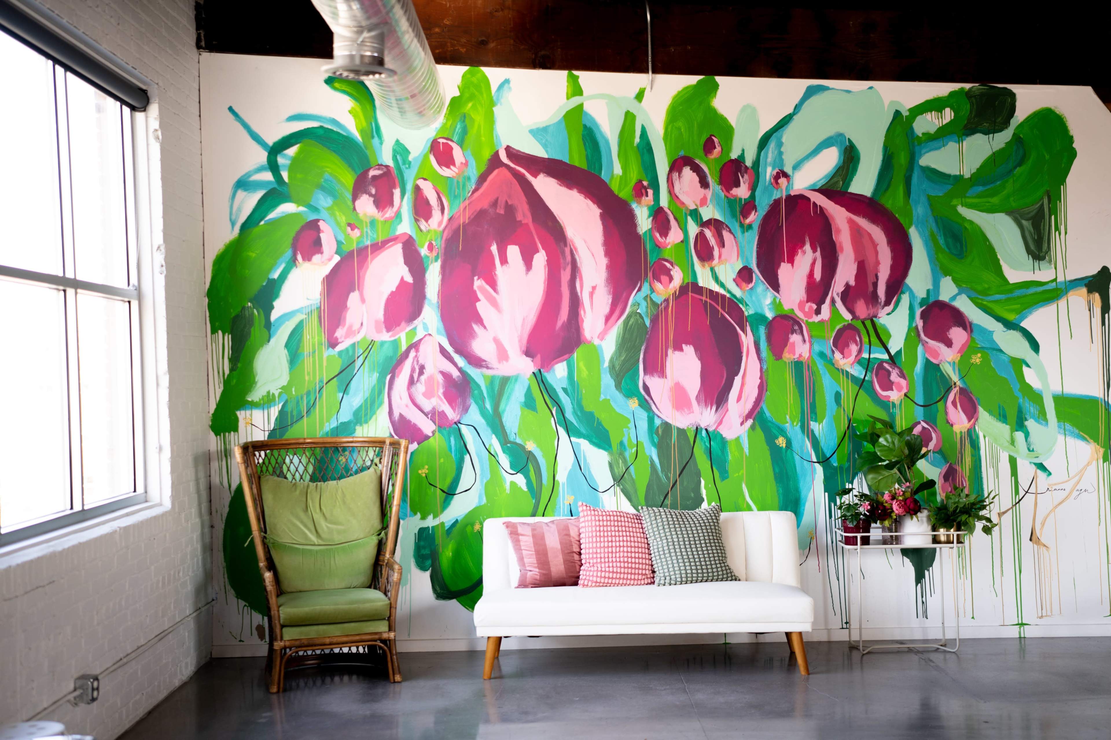 The image features a bright interior with a large floral mural on the wall, a white sofa, a colorful pillow arrangement, and a green chair.