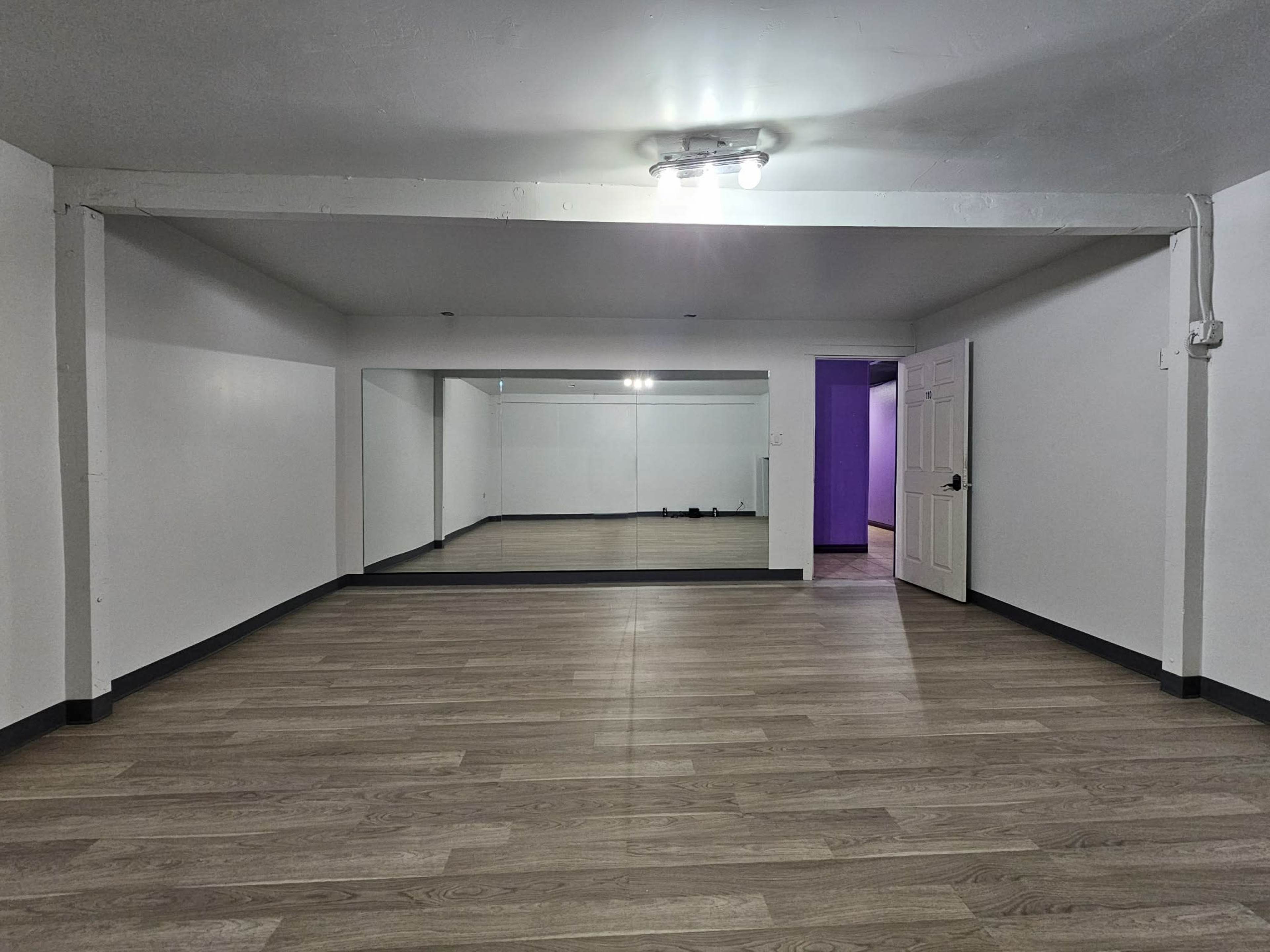 The image shows a spacious, empty room with wooden flooring, white walls, and a large mirror on one side.