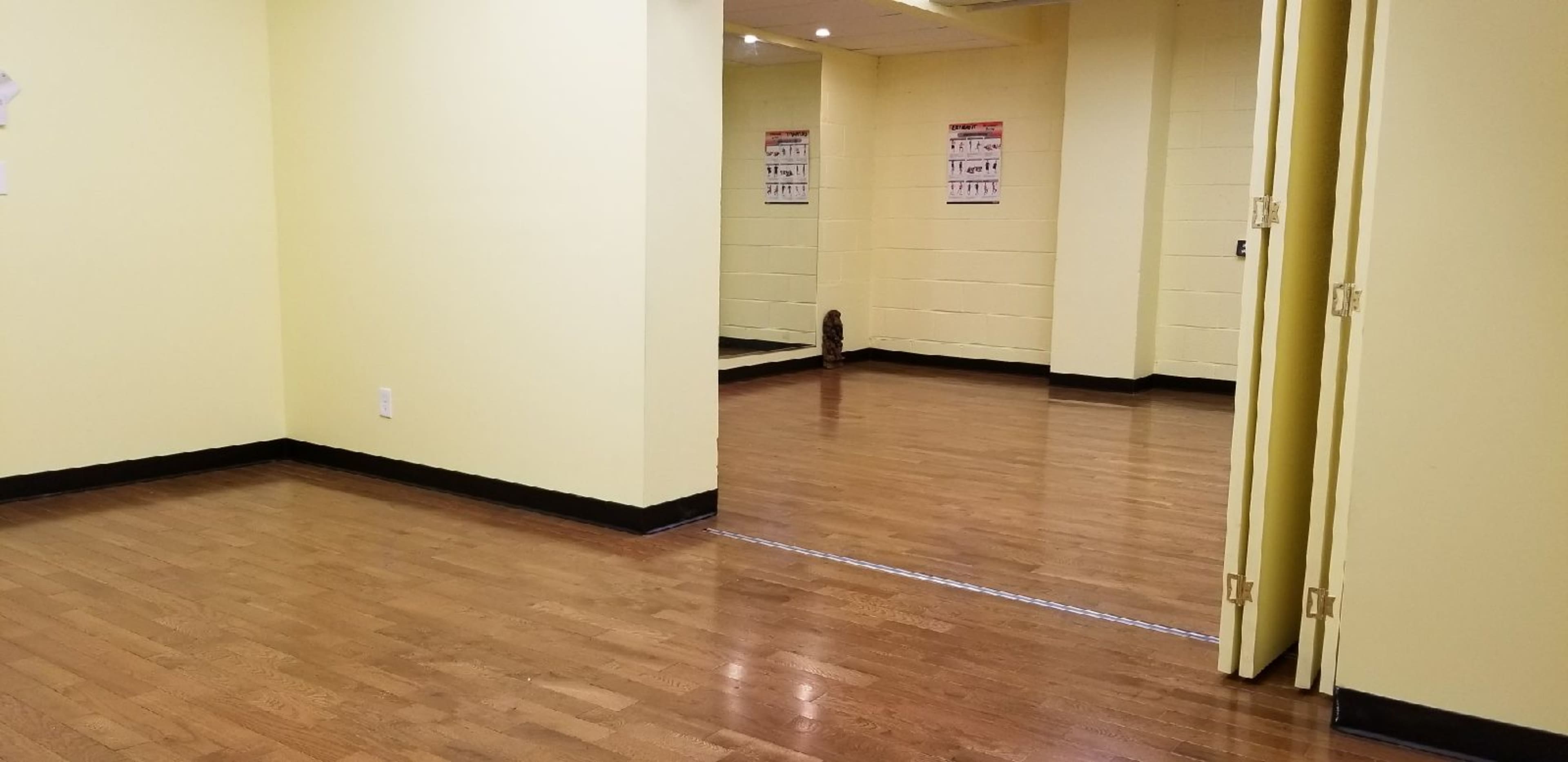 The image shows a bright, empty room with yellow walls and wooden flooring, featuring a mirrored wall and two doorways.