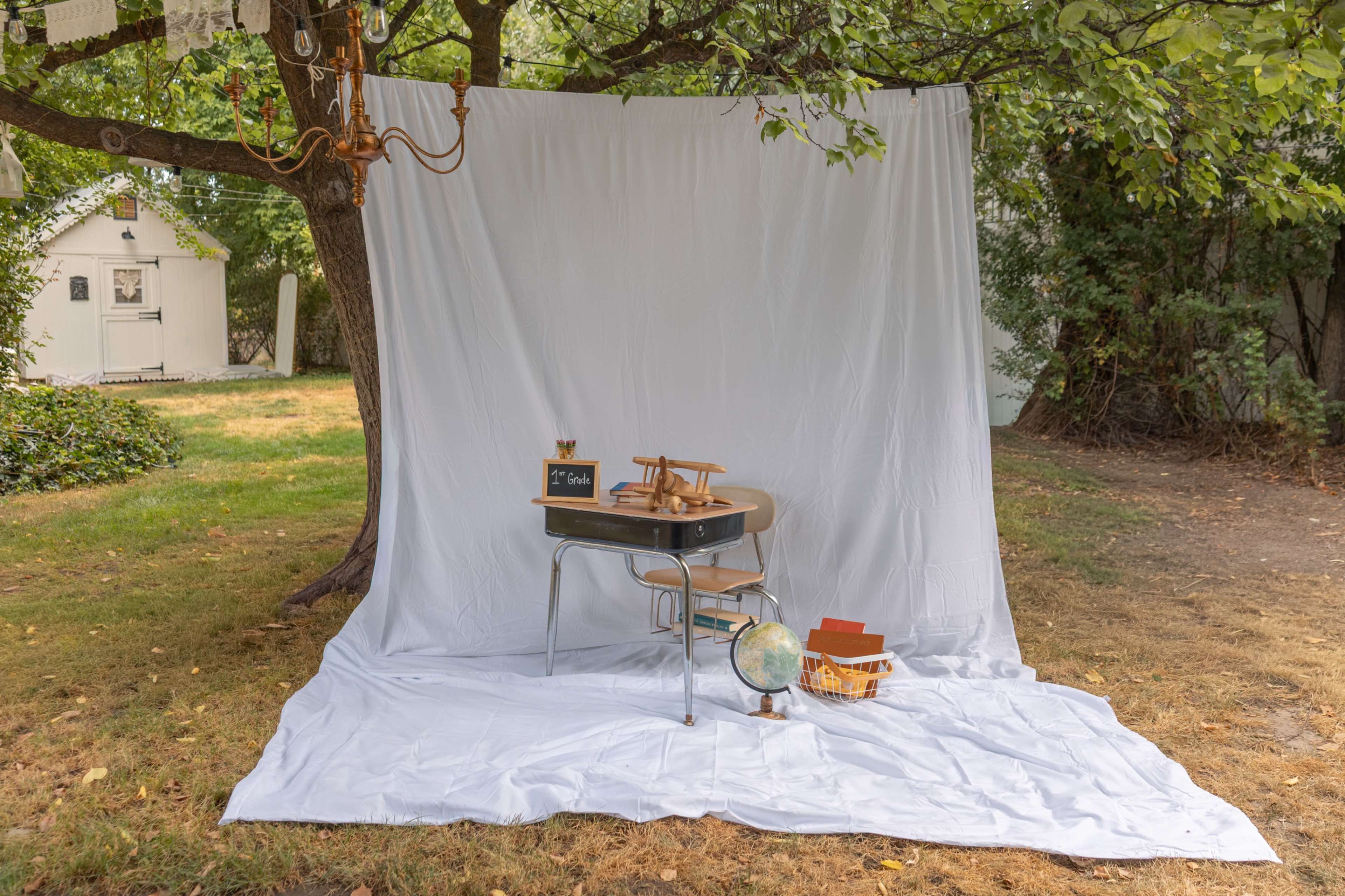 A simple outdoor setup features a white backdrop draped from a tree, a desk with wooden chairs, a globe, and a basket of books on a grassy area.