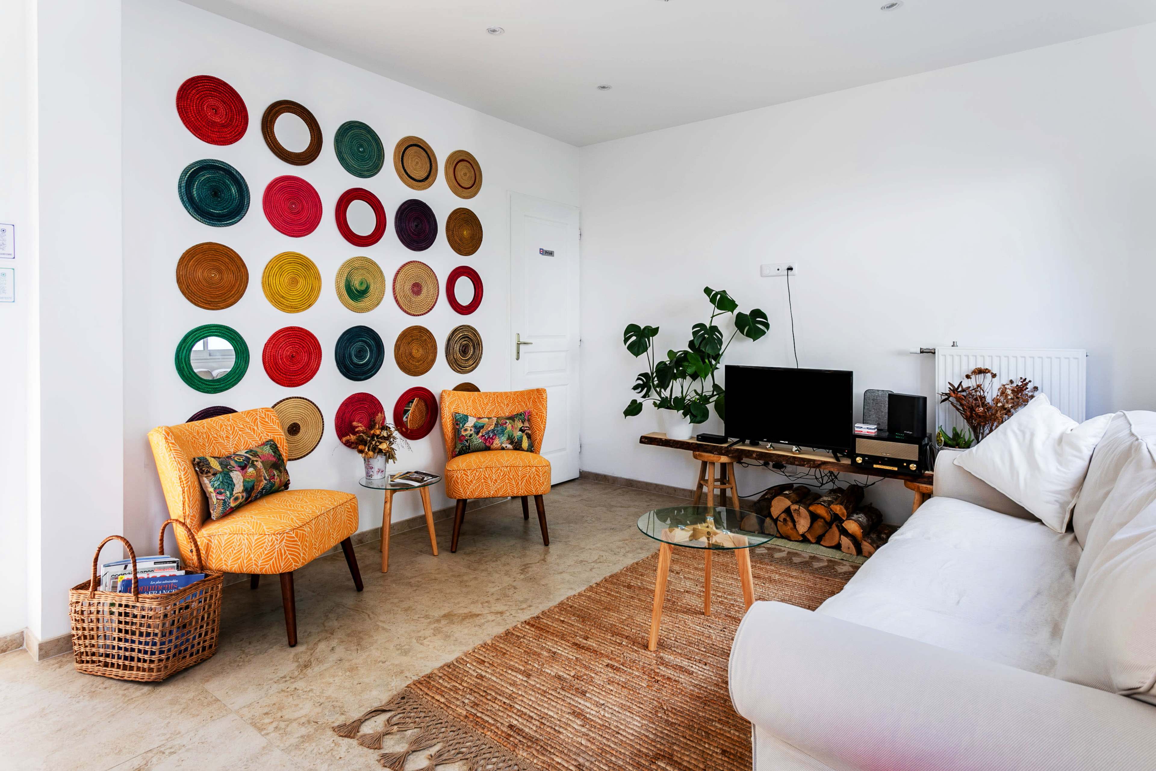 A bright living room with two orange chairs, a white sofa, a coffee table, a television, and a wall adorned with colorful decorative plates.