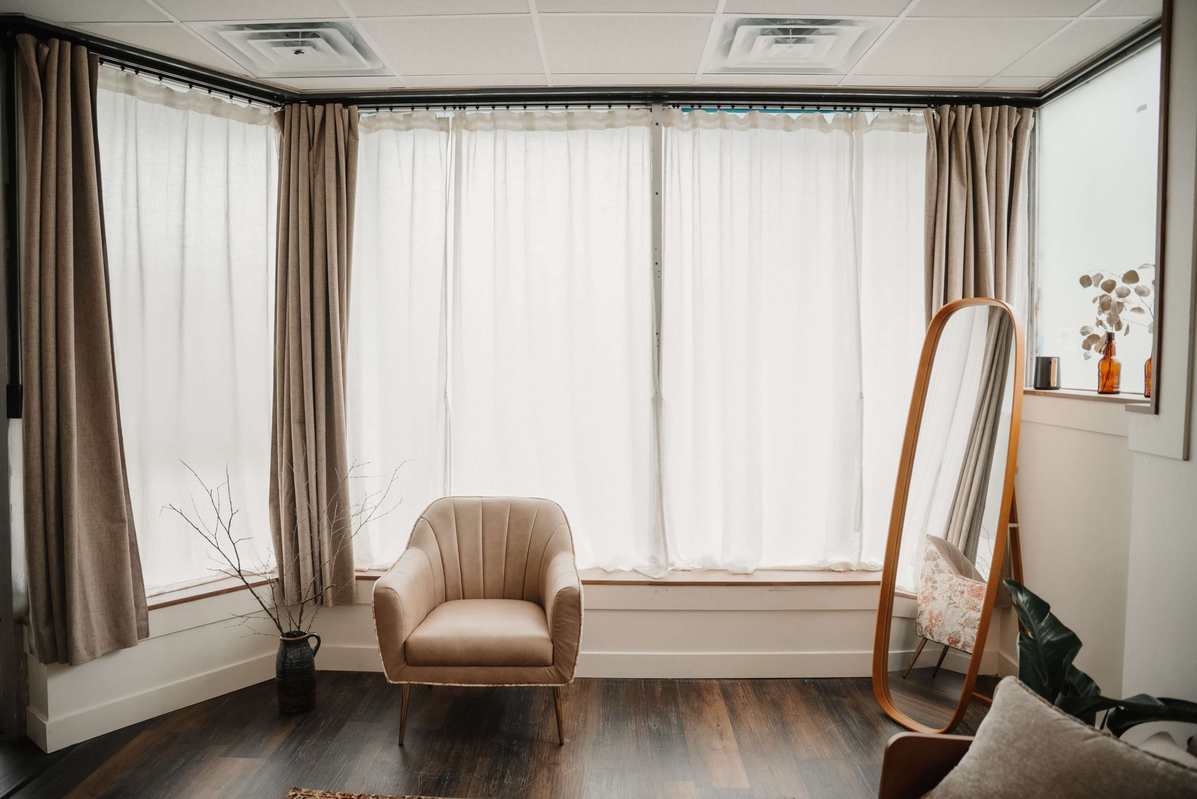A cozy corner features a beige armchair next to a large mirror and light curtains, set against a wooden floor.