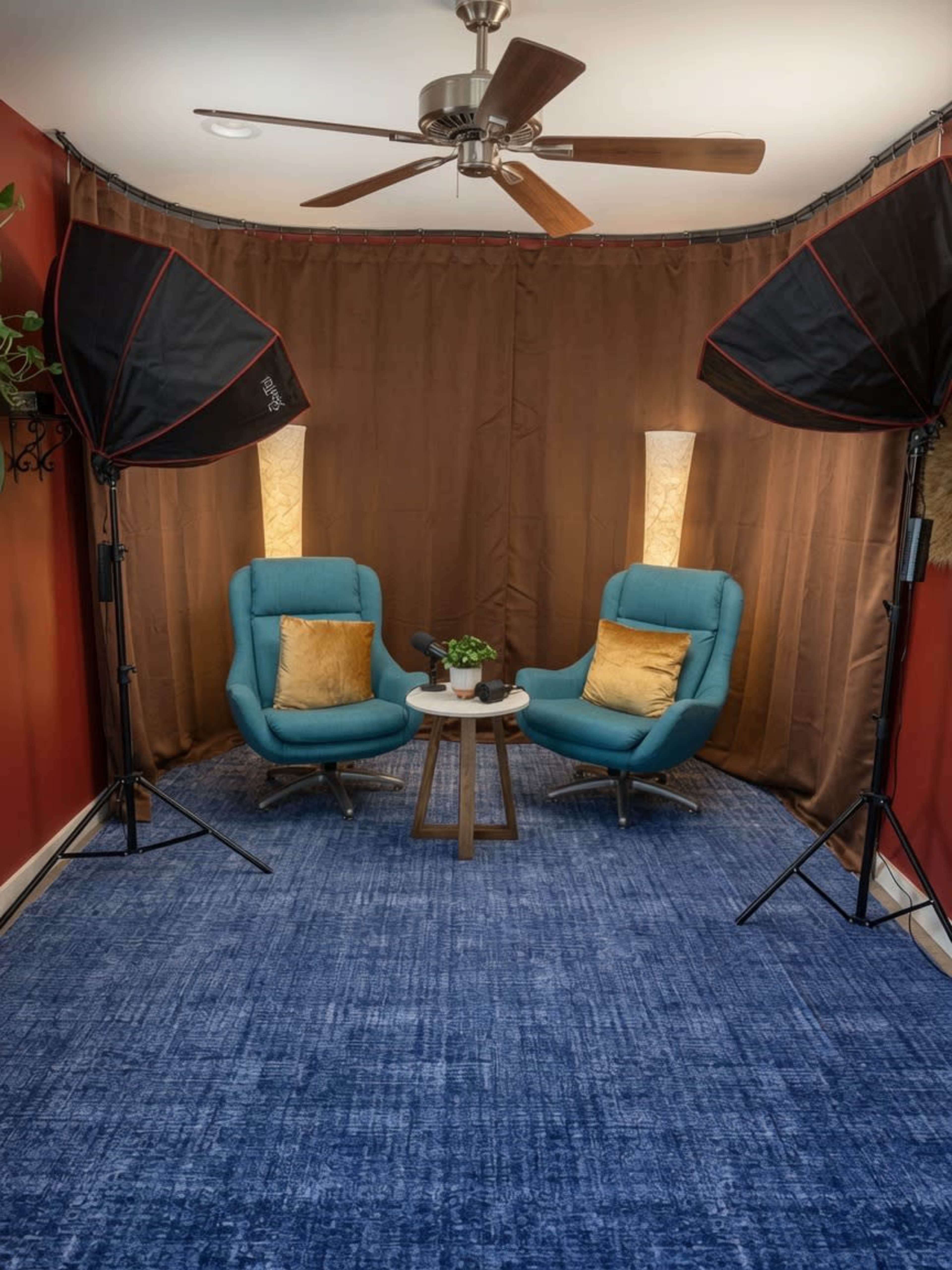 The image shows a cozy studio setup with two turquoise armchairs, a small round table, and soft lighting, all framed by brown curtains.