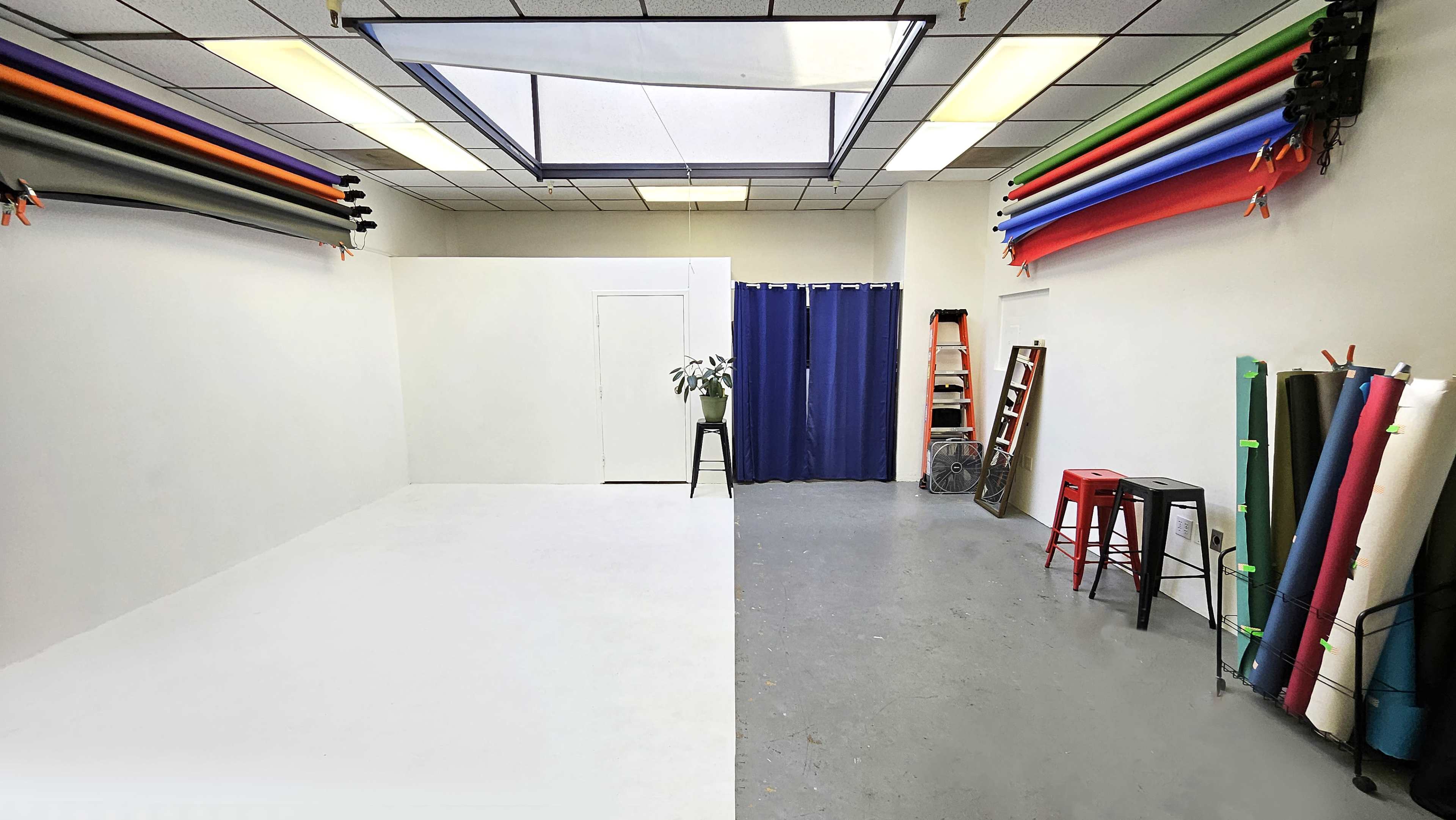 A studio space with a white backdrop, a large skylight, colorful backdrop rolls on the walls, and a few stools and a ladder positioned in the room.