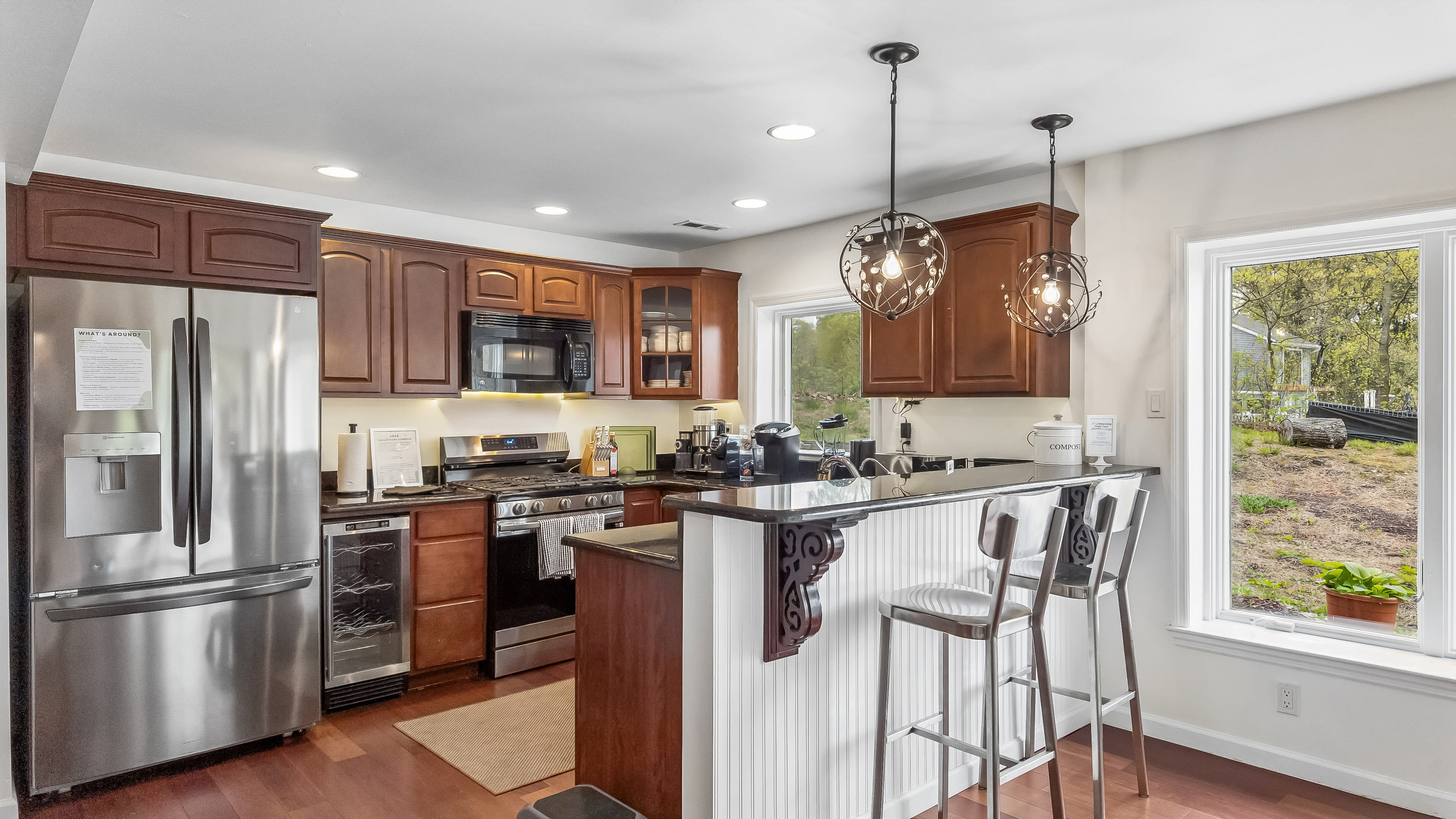 The image shows a modern kitchen with wood cabinetry, stainless steel appliances, and a breakfast bar with two stools.