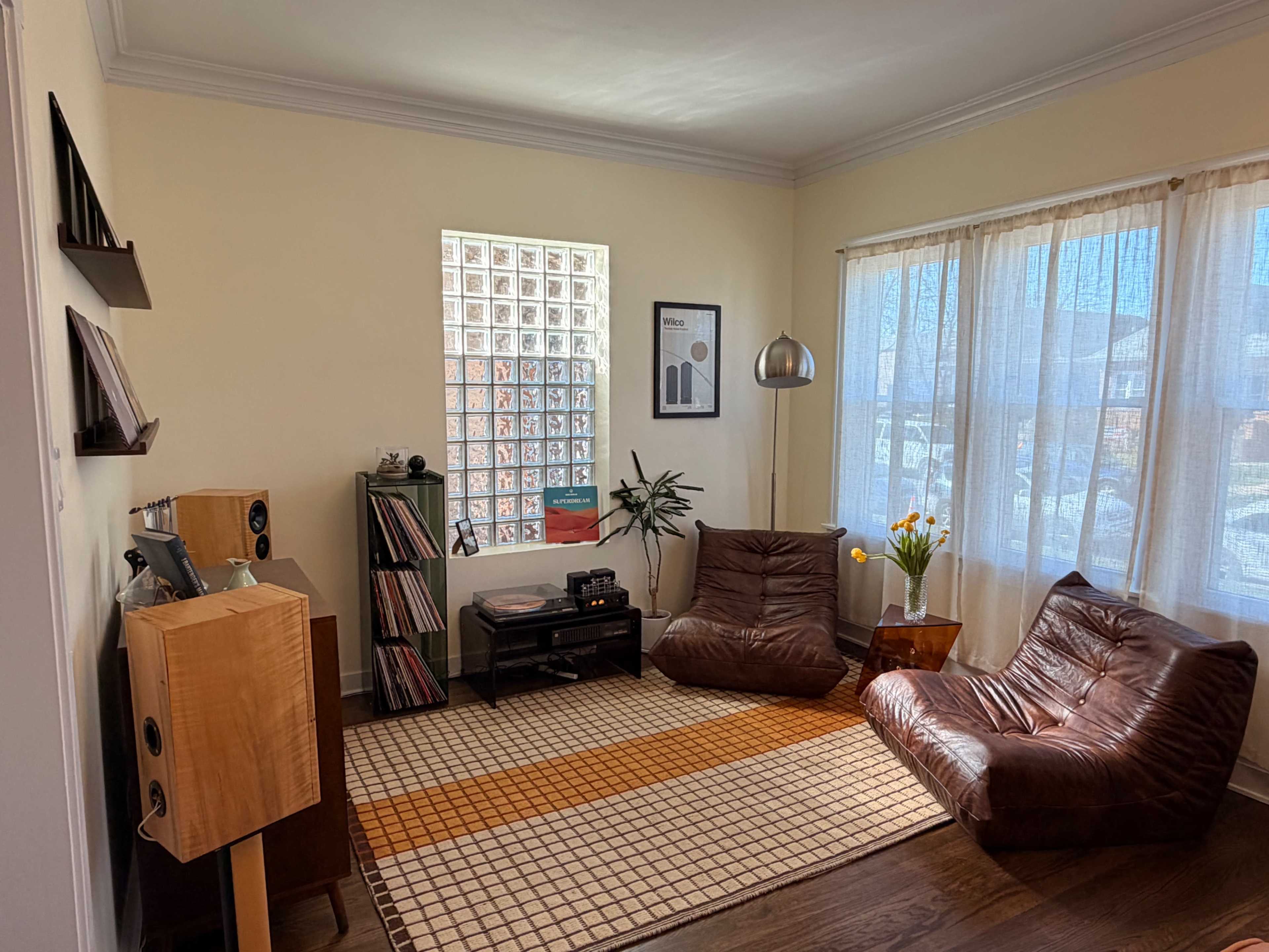 The image shows a well-lit living room with two brown leather chairs, a wooden speaker, a record player, and a window featuring decorative glass blocks.