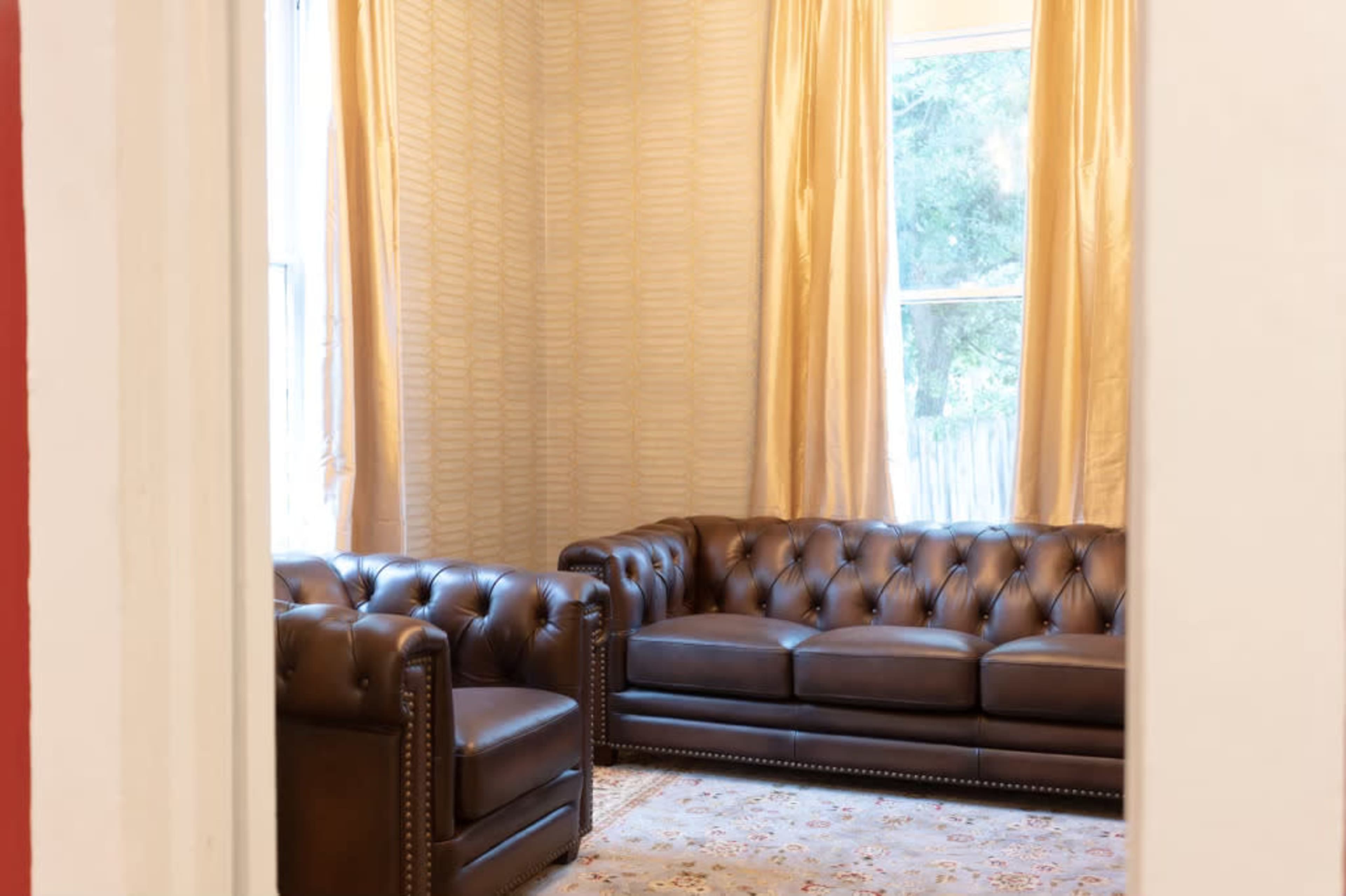 A room with a leather chesterfield sofa and an armchair, with gold curtains framing a window in the background.