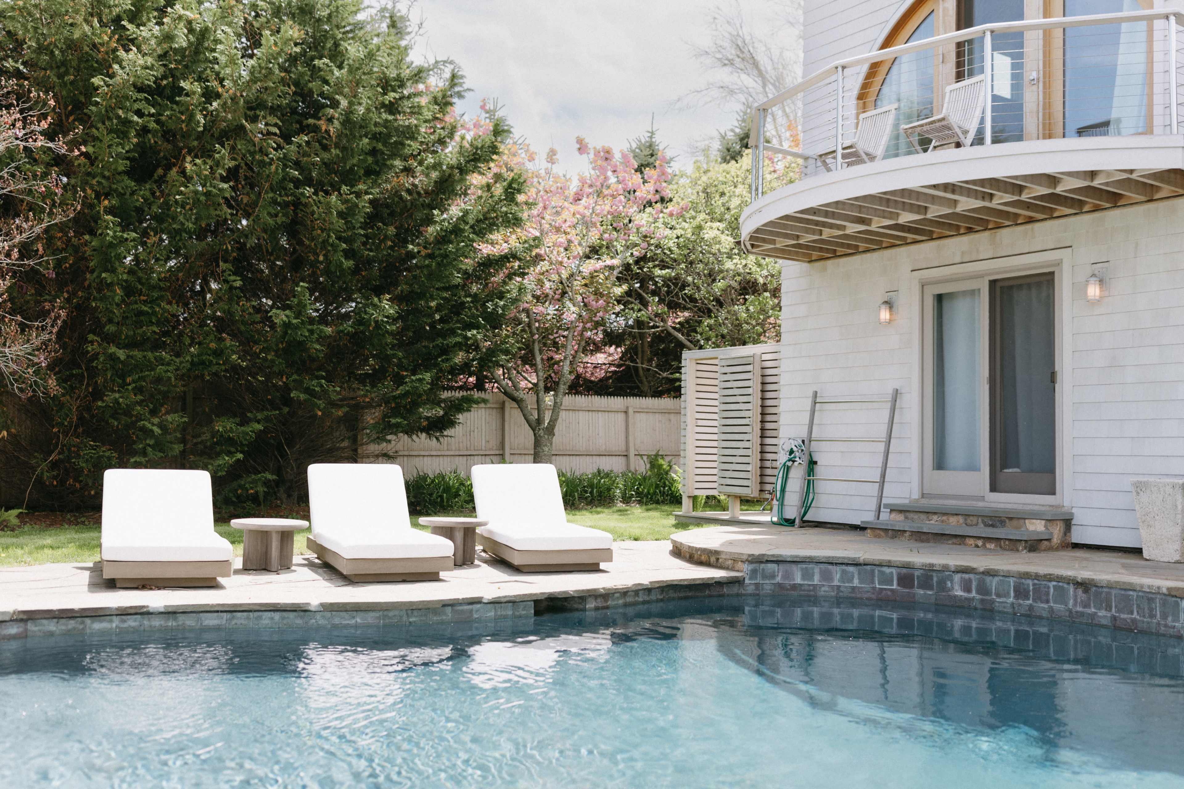 The image features a serene backyard scene with a swimming pool, three white lounge chairs, and a modern house surrounded by greenery.