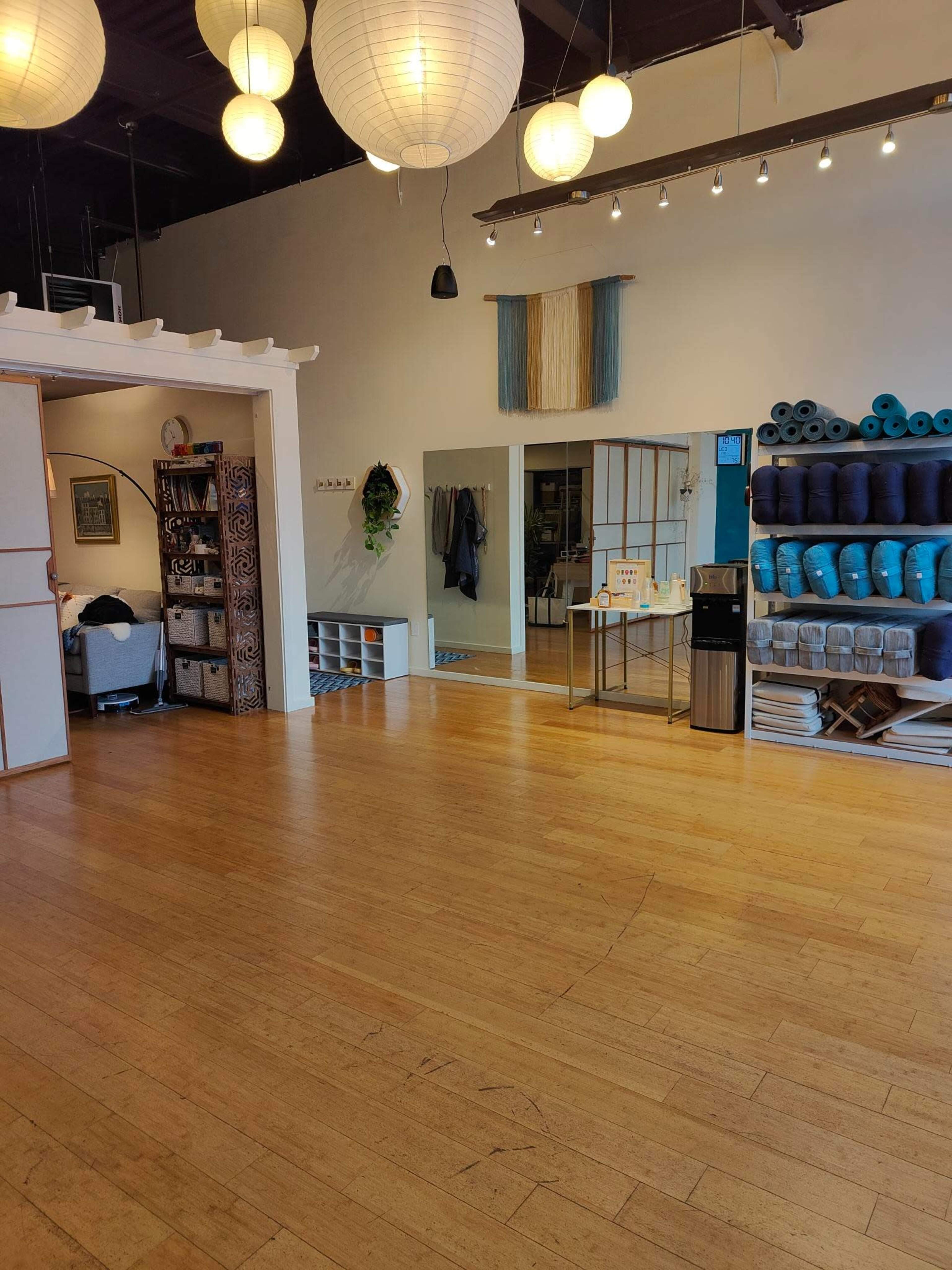 A spacious interior of a studio with wooden floors, decorative paper lanterns, and organized shelves displaying yoga mats and towels.