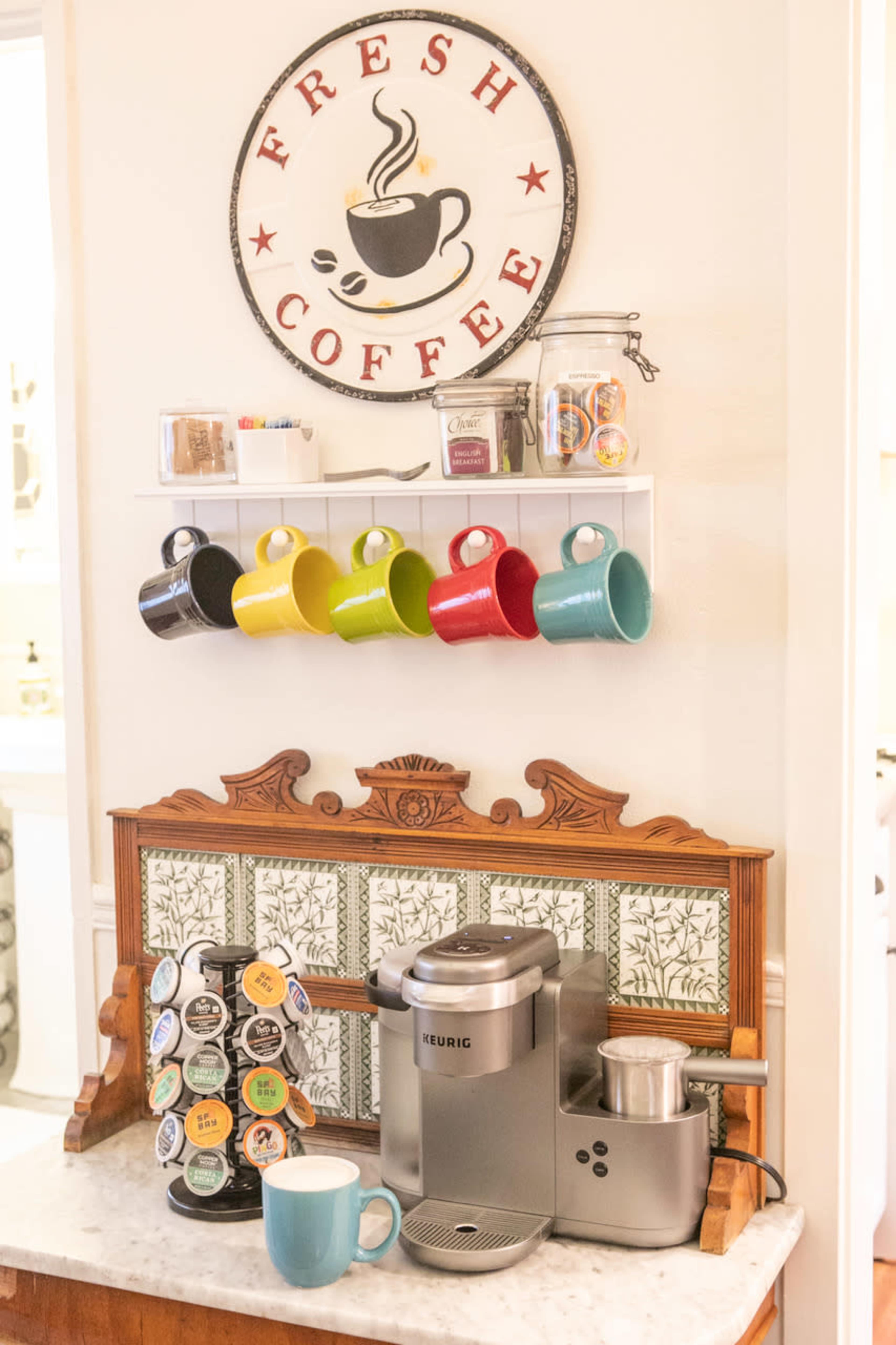 A coffee station features a Keurig machine, a variety of coffee pods, and multicolored mugs displayed on a shelf, all beneath a circular "Fresh Coffee" sign.