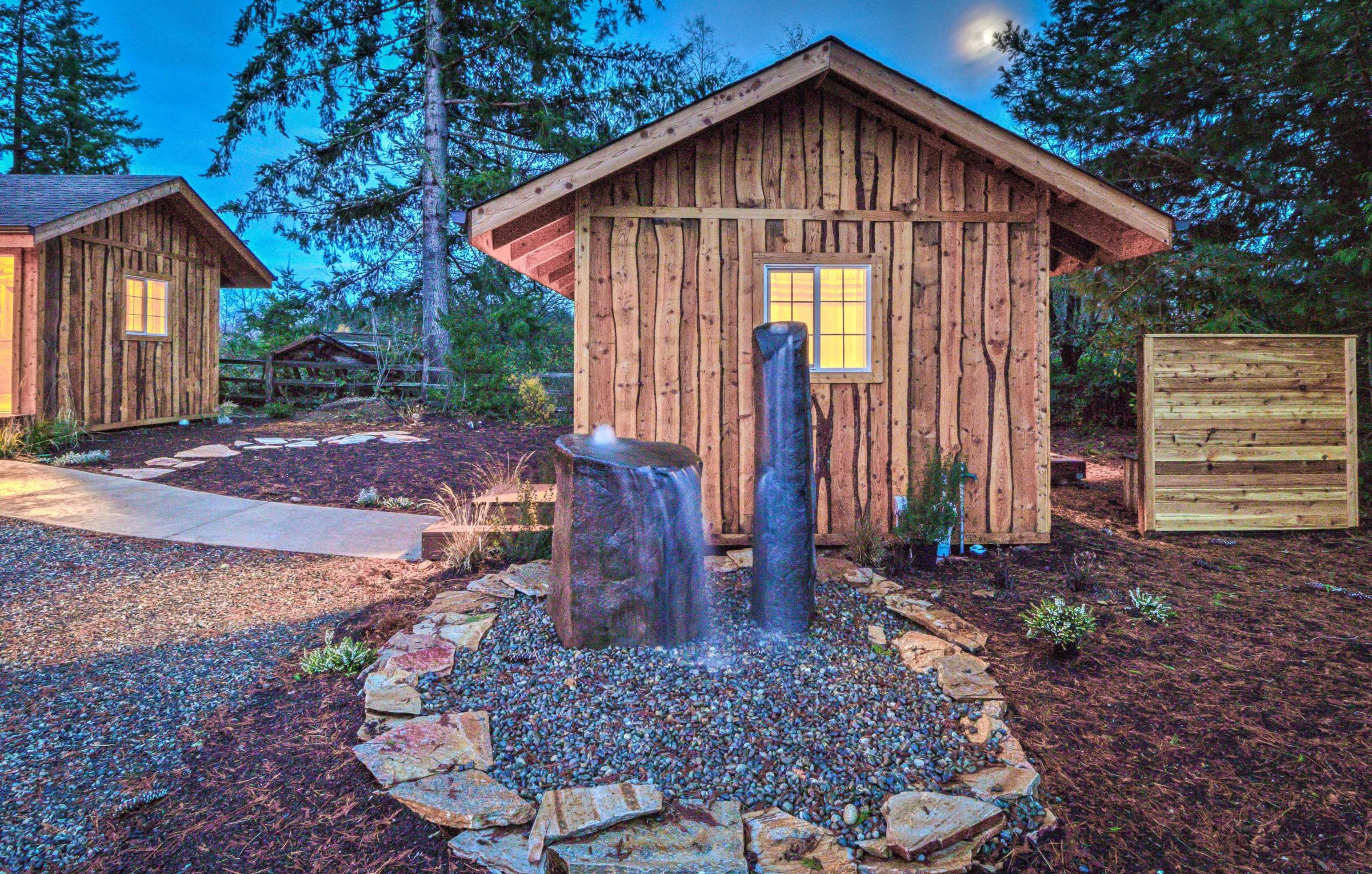 A wooden cabin surrounded by a stone path, a small water feature, and landscaped greenery under a dusk sky.