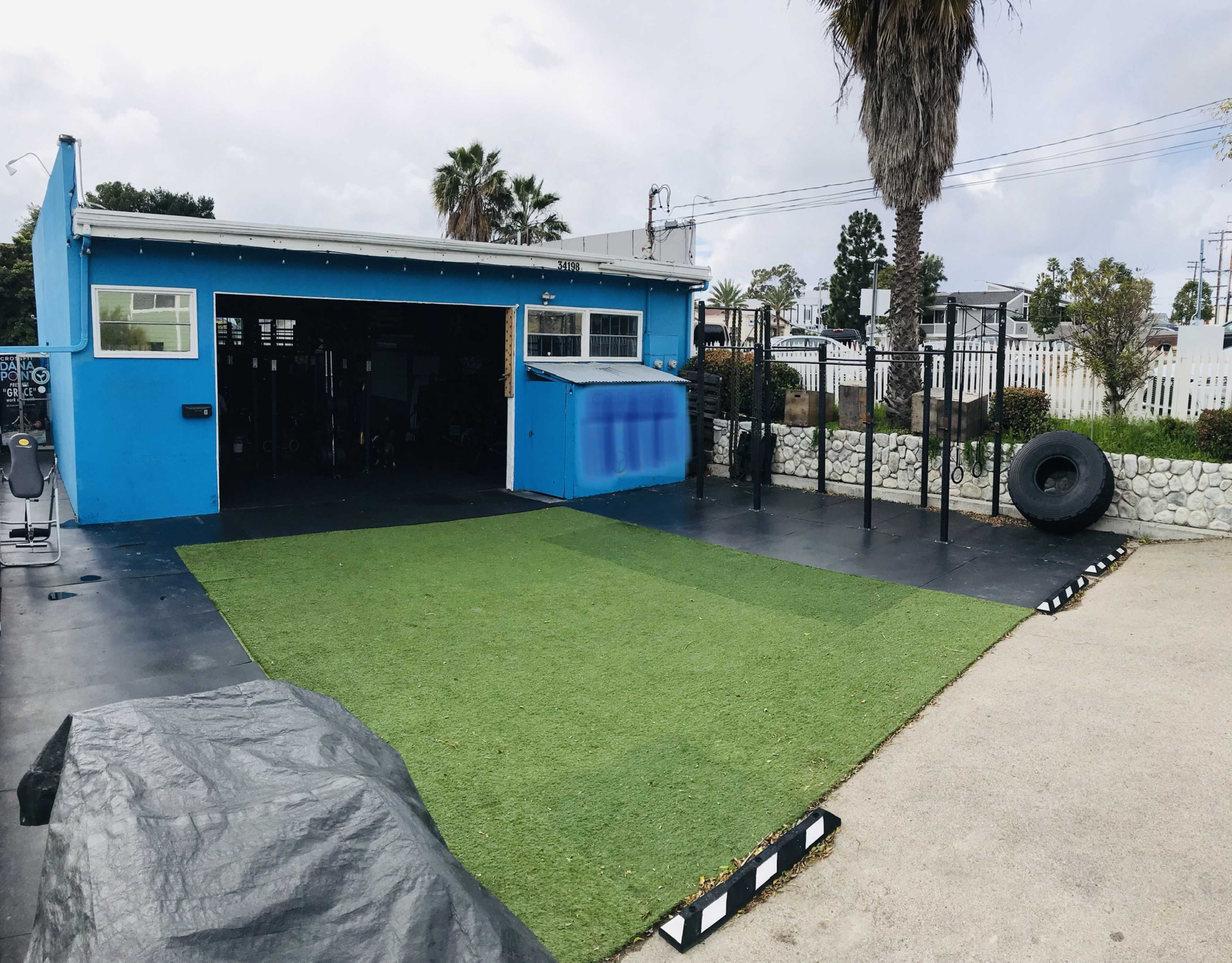 The image shows an outdoor gym area featuring turf flooring, exercise equipment, and a blue building in the background.