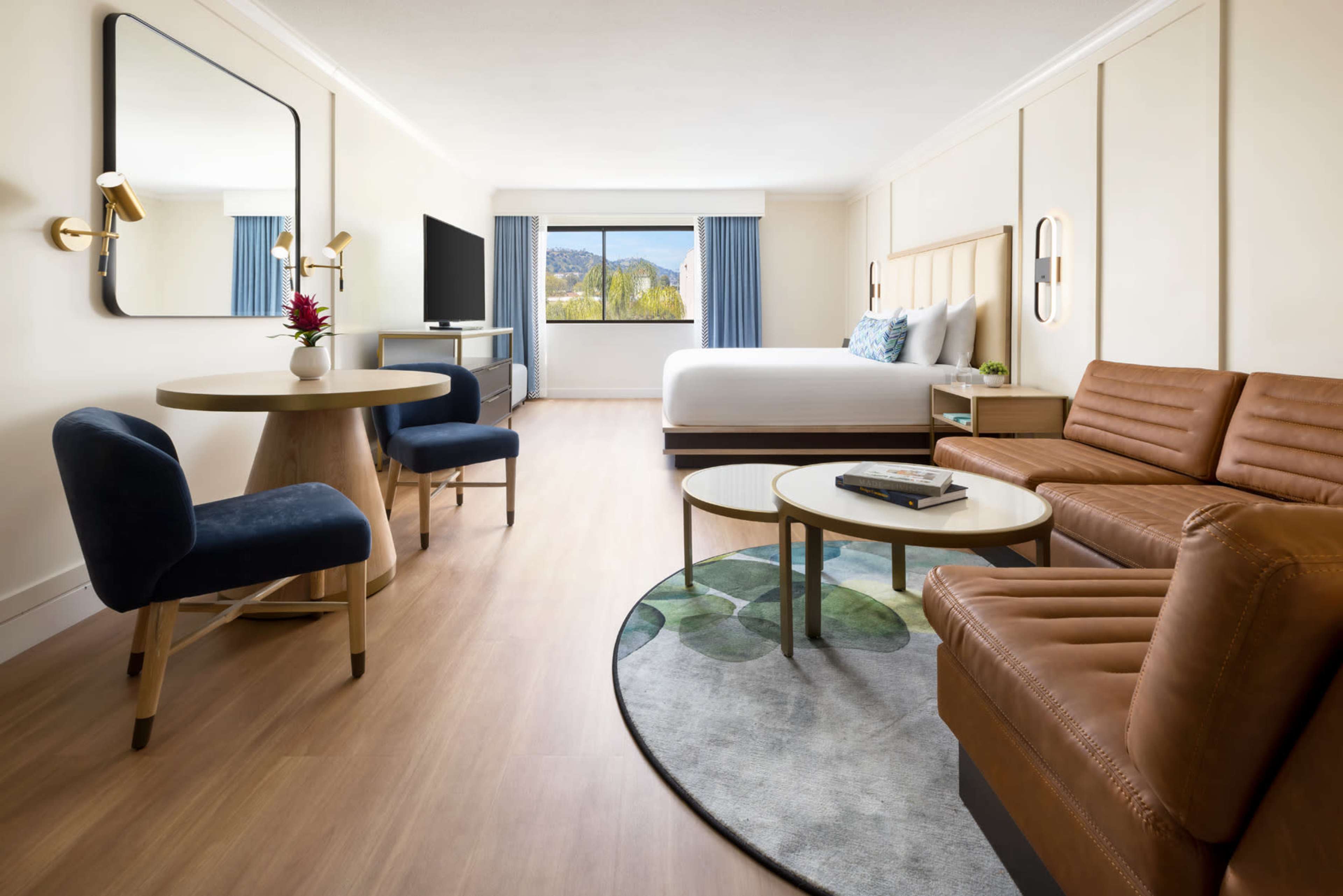 The image depicts a modern hotel room featuring a bed, seating area with a brown leather sofa, a table with chairs, and a large window with a view.