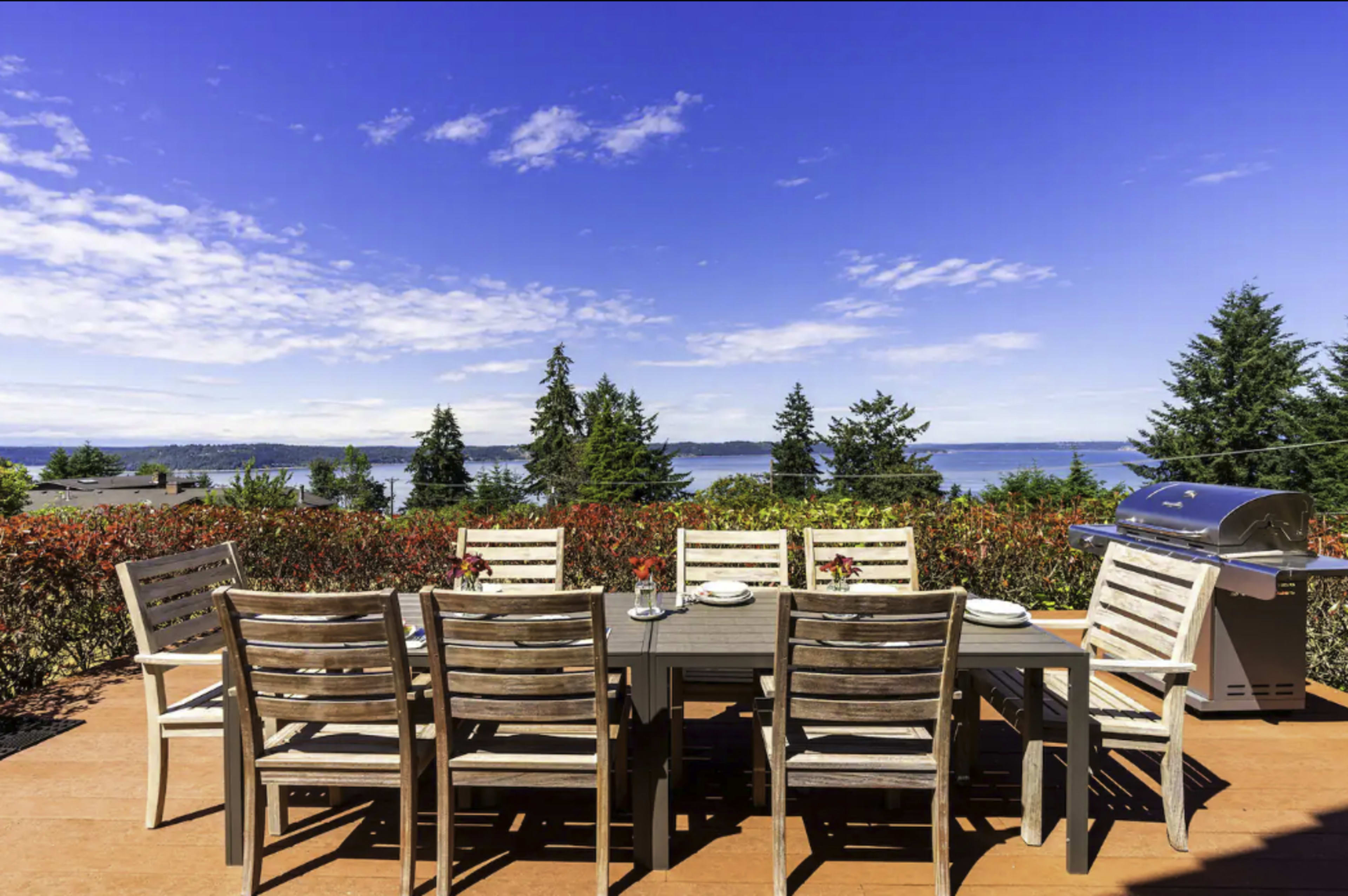 A wooden dining table is set with plates and glasses, surrounded by eight chairs, overlooking a landscape of trees and a body of water.