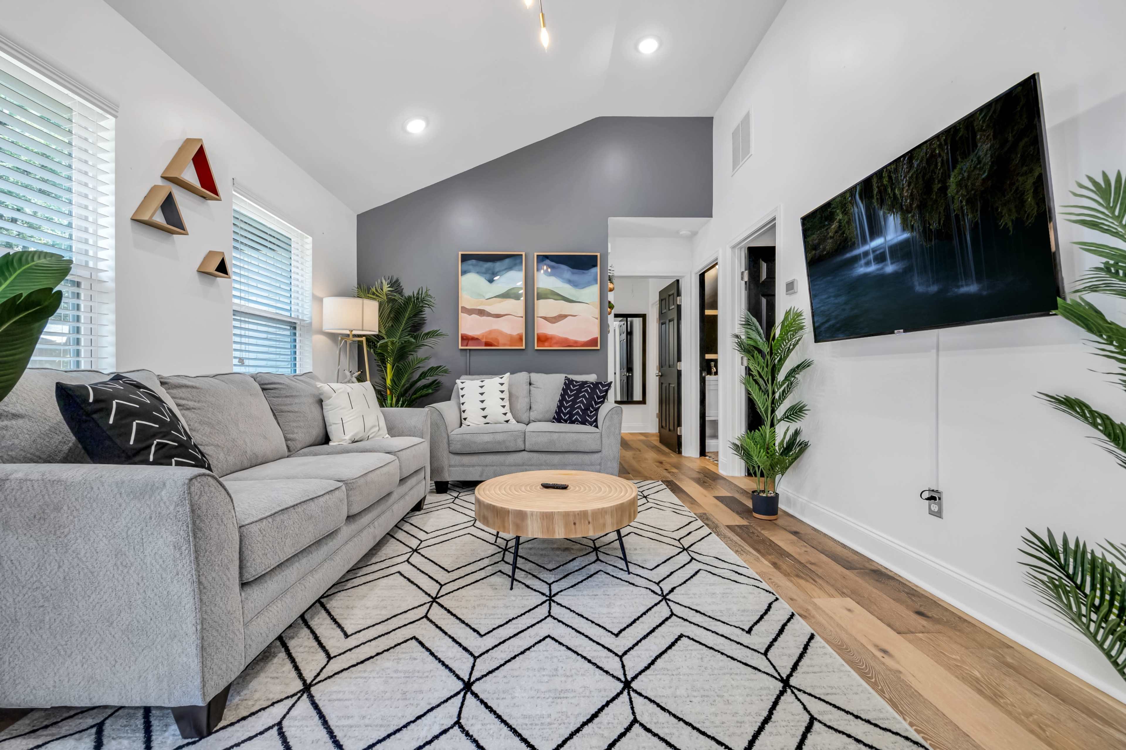 A modern living room with a gray sofa, a round wooden coffee table, vibrant artwork on the wall, and lush houseplants.