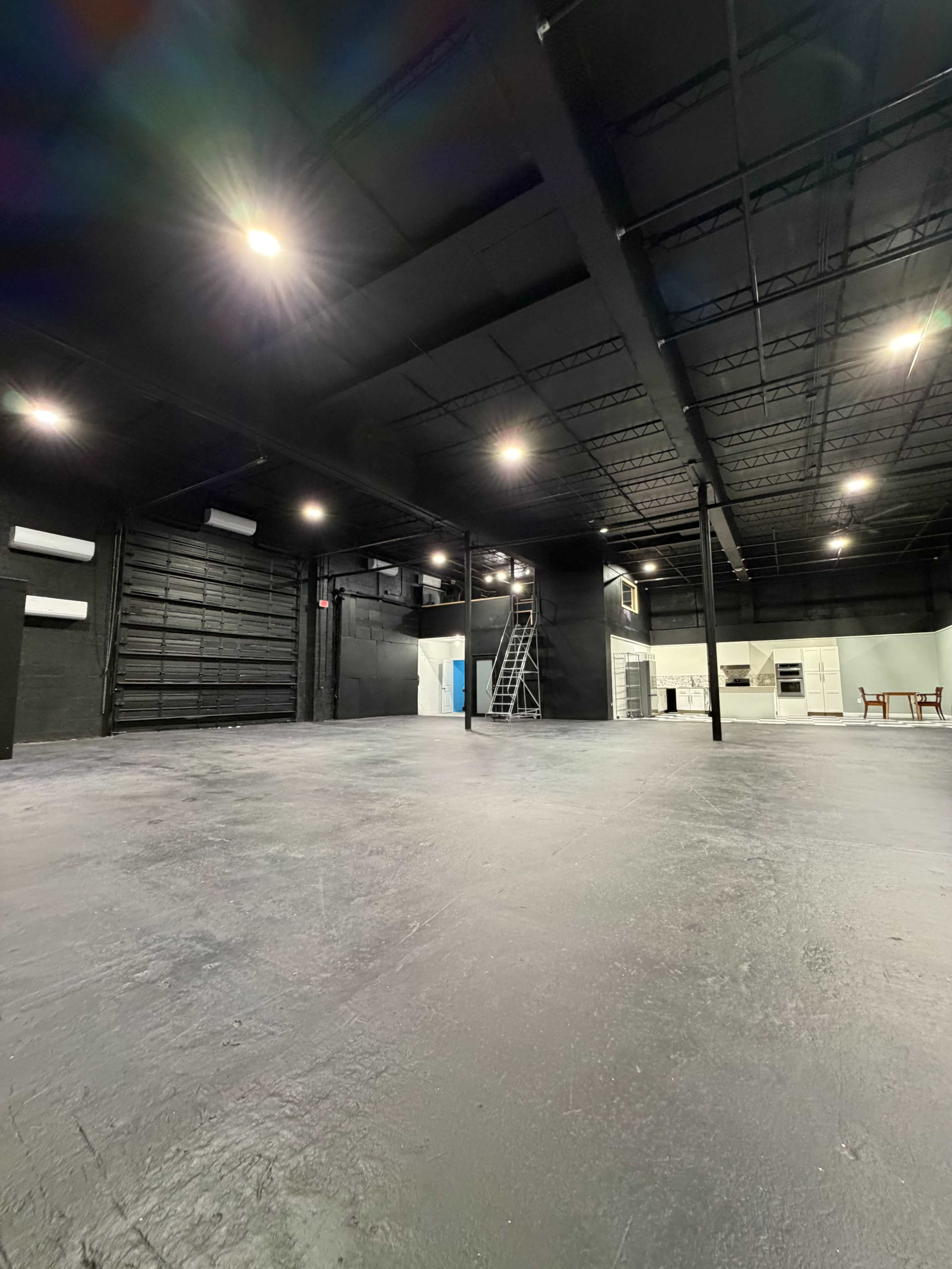 The image depicts a large, empty industrial space with a dark ceiling and bright overhead lighting, featuring a smooth floor and minimalist decor.