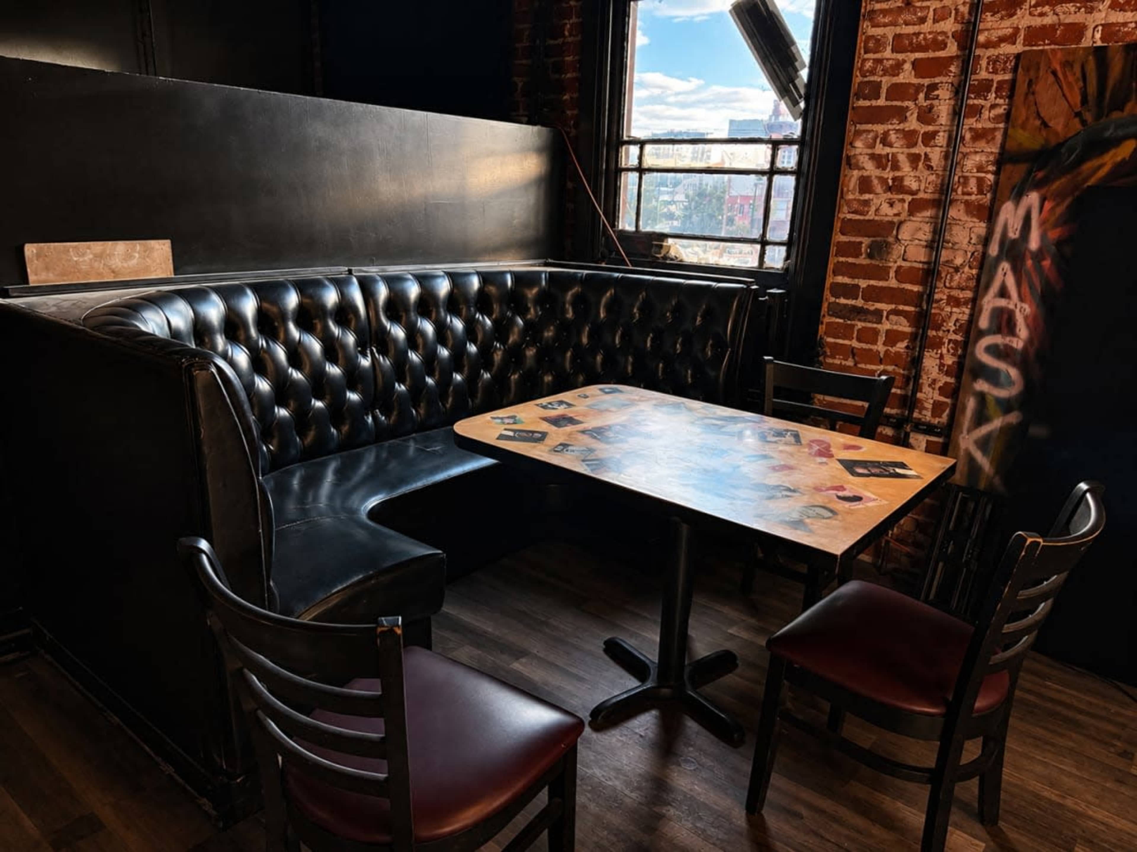 The image shows a cozy, leather-upholstered booth with a wooden table and several wooden chairs in an industrial-style interior featuring exposed brick walls.