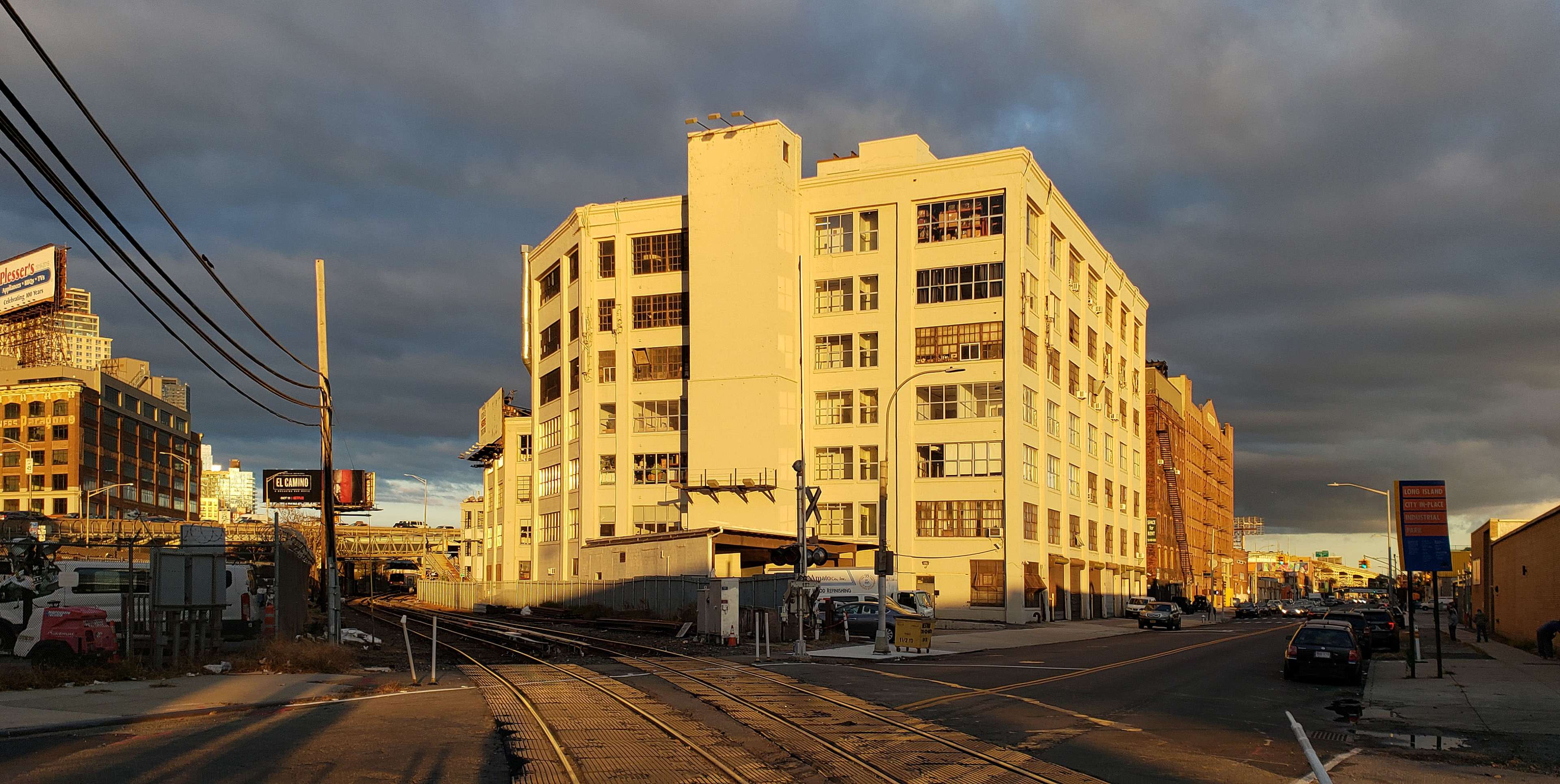 A white, five-story industrial building stands next to a set of railway tracks under a cloudy sky.