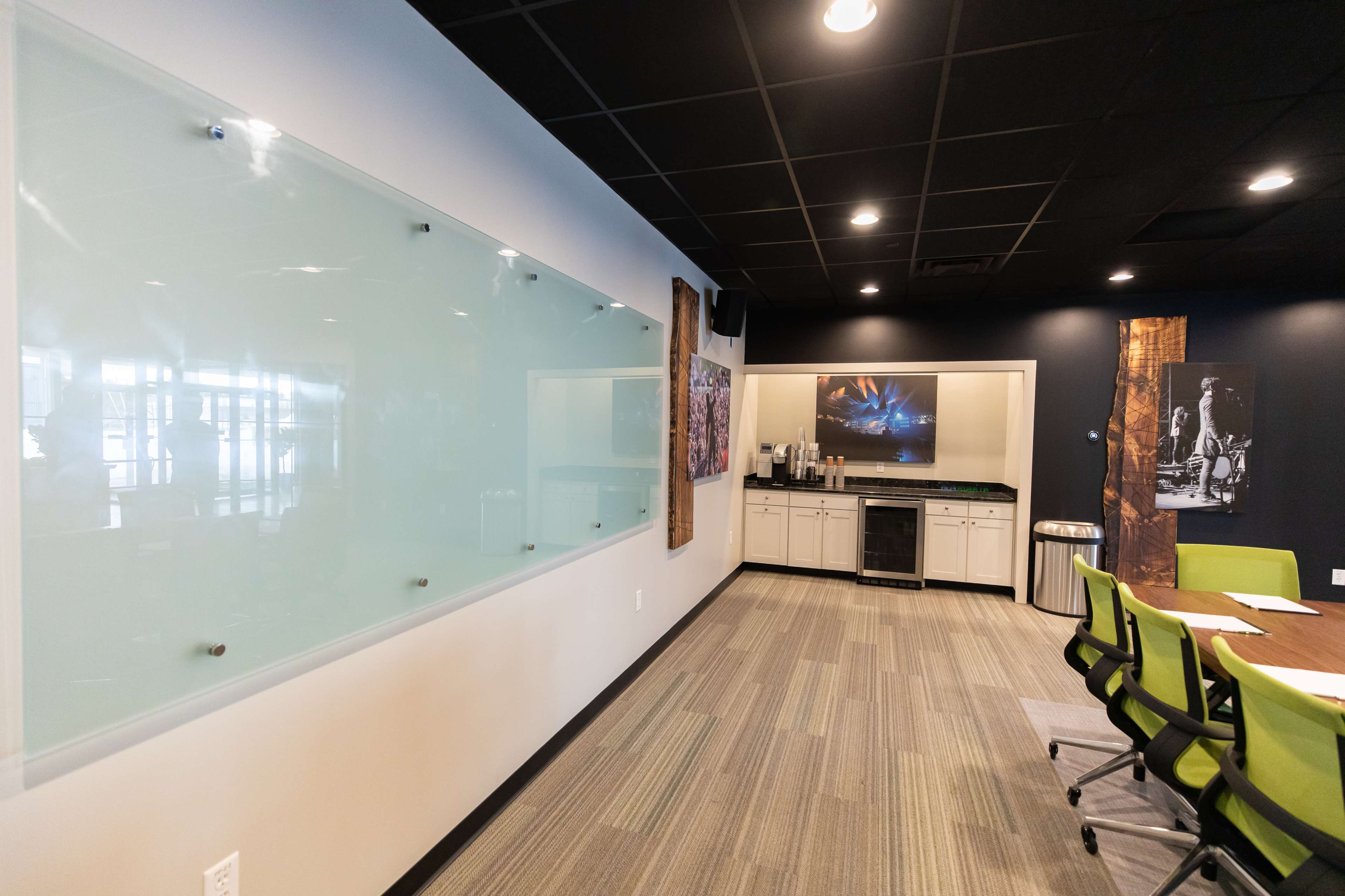 A modern conference room with a glass whiteboard, a kitchenette area, and a large table surrounded by green chairs.