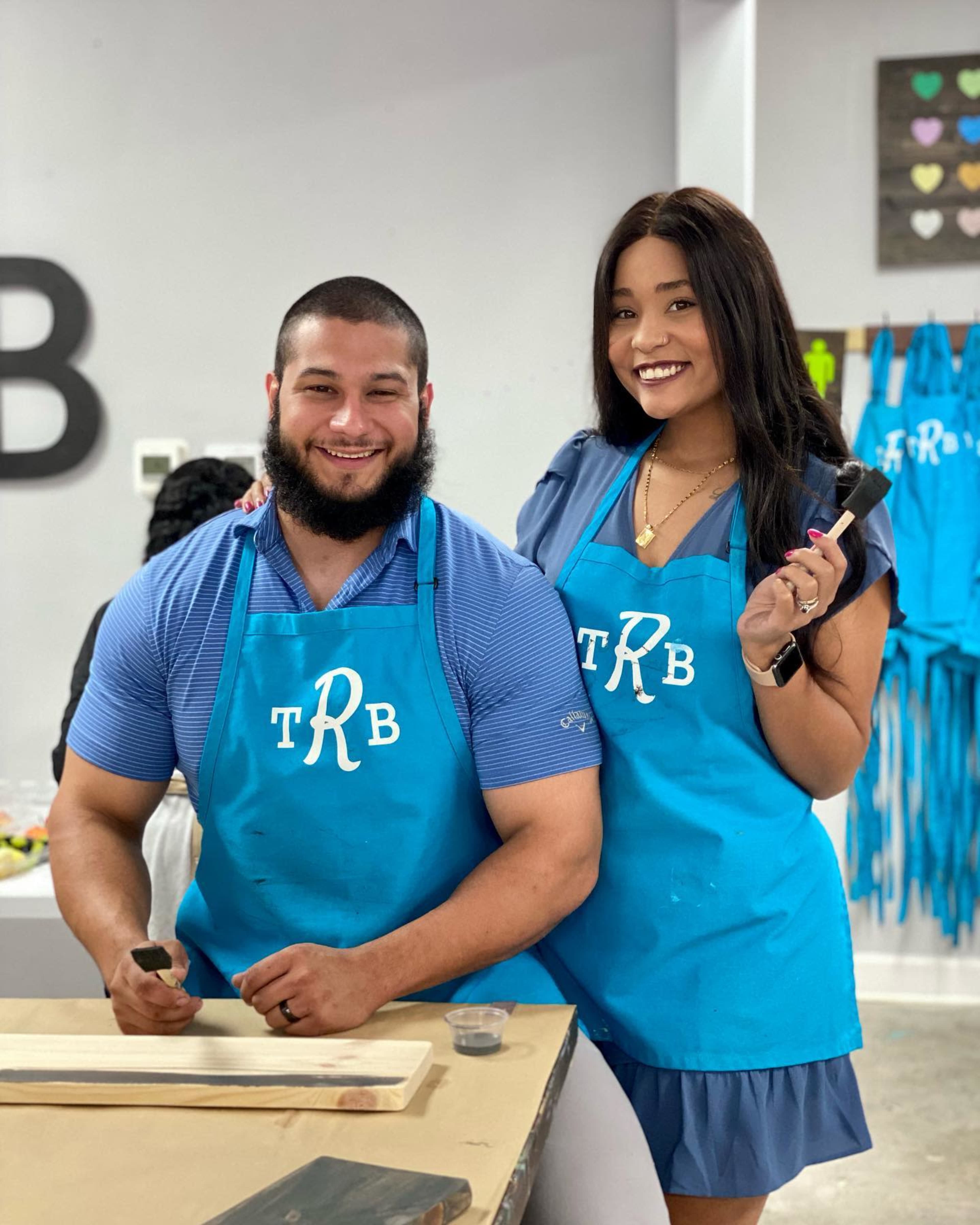 A man and a woman wearing matching blue aprons pose together in a workshop setting.