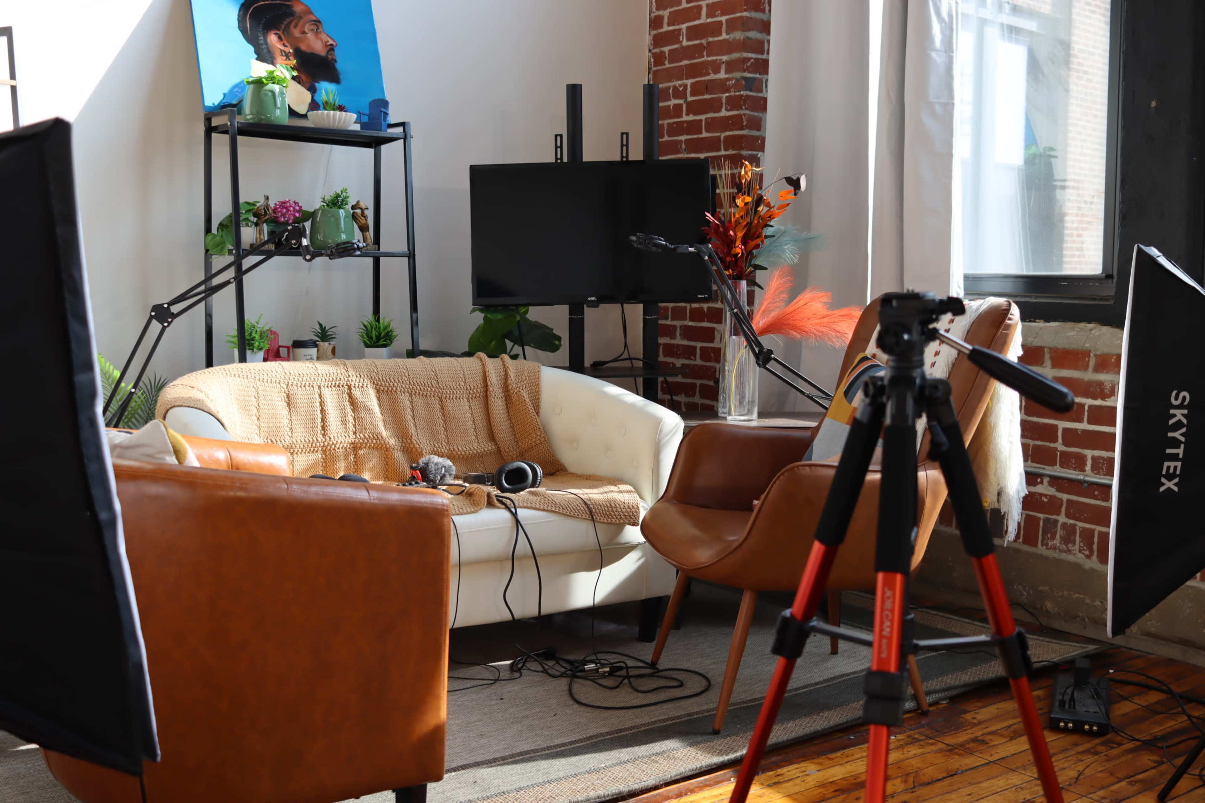 A modern living space with a beige couch, two brown chairs, and a camera tripod set up in front of a television against a backdrop of exposed brick walls.