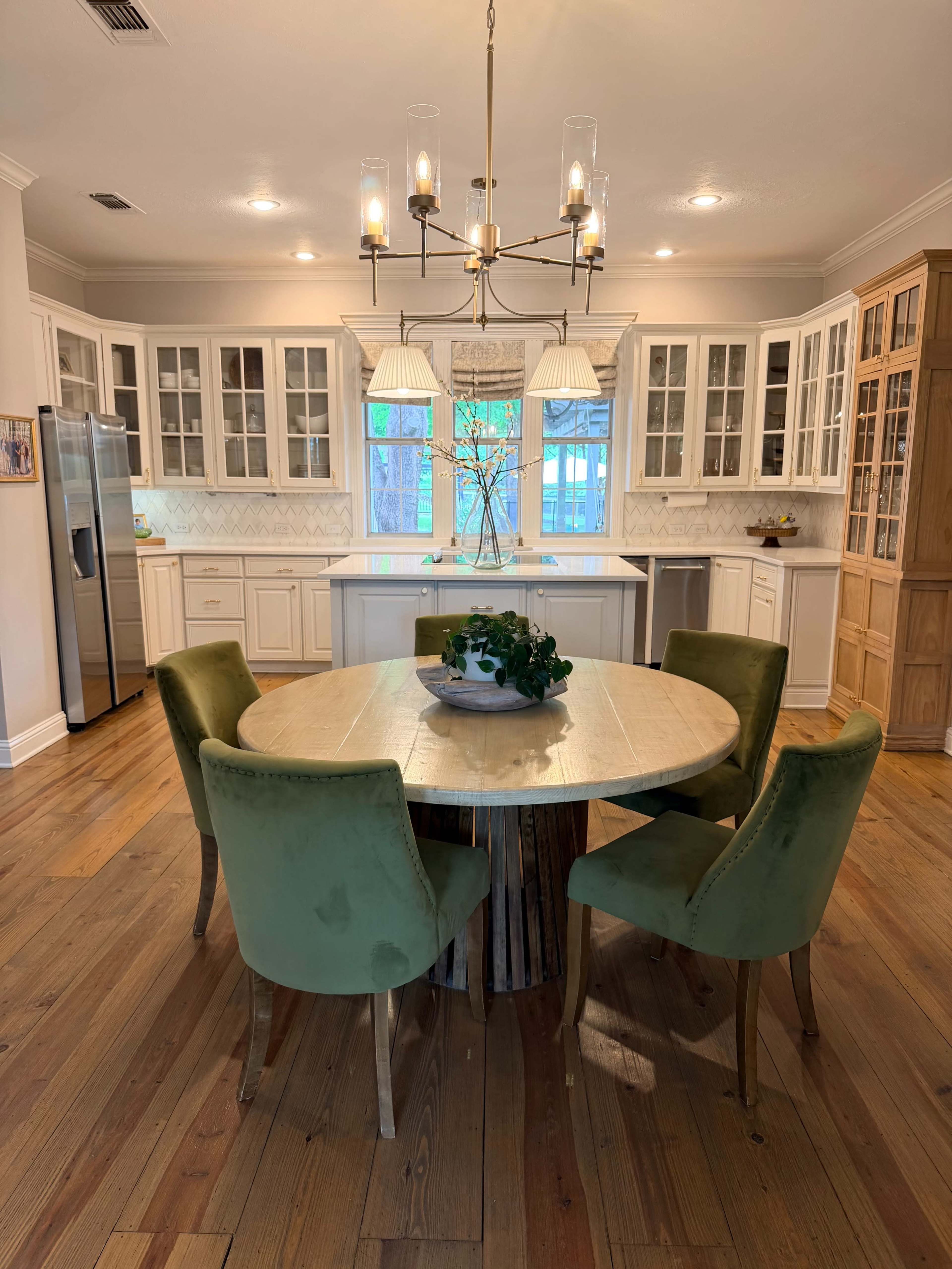 A round wooden dining table surrounded by green upholstered chairs is positioned in a spacious kitchen with white cabinetry and windows allowing natural light.