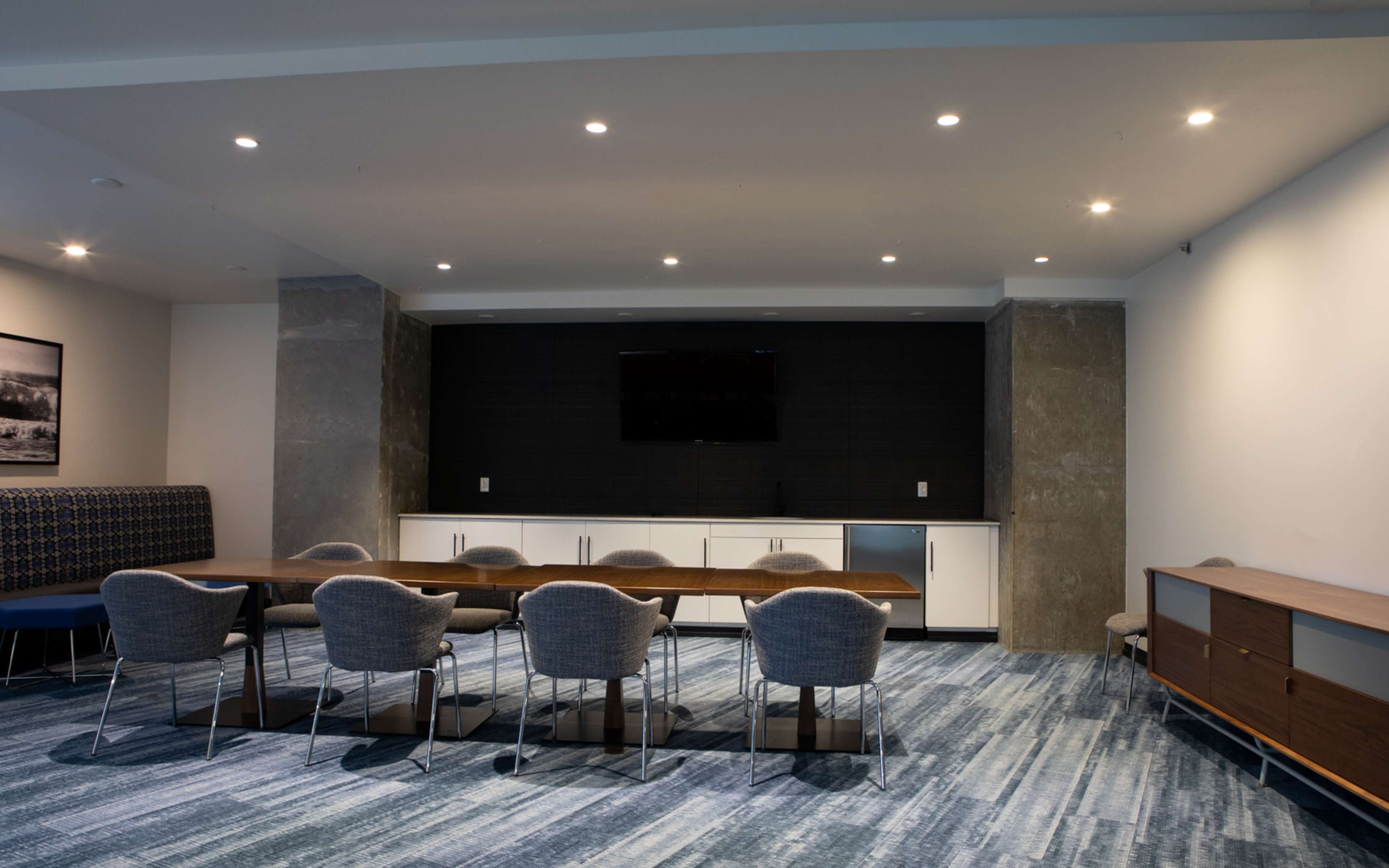 A modern meeting room with a long wooden table surrounded by upholstered chairs, a wall-mounted television, and a side cabinet against a textured wall.