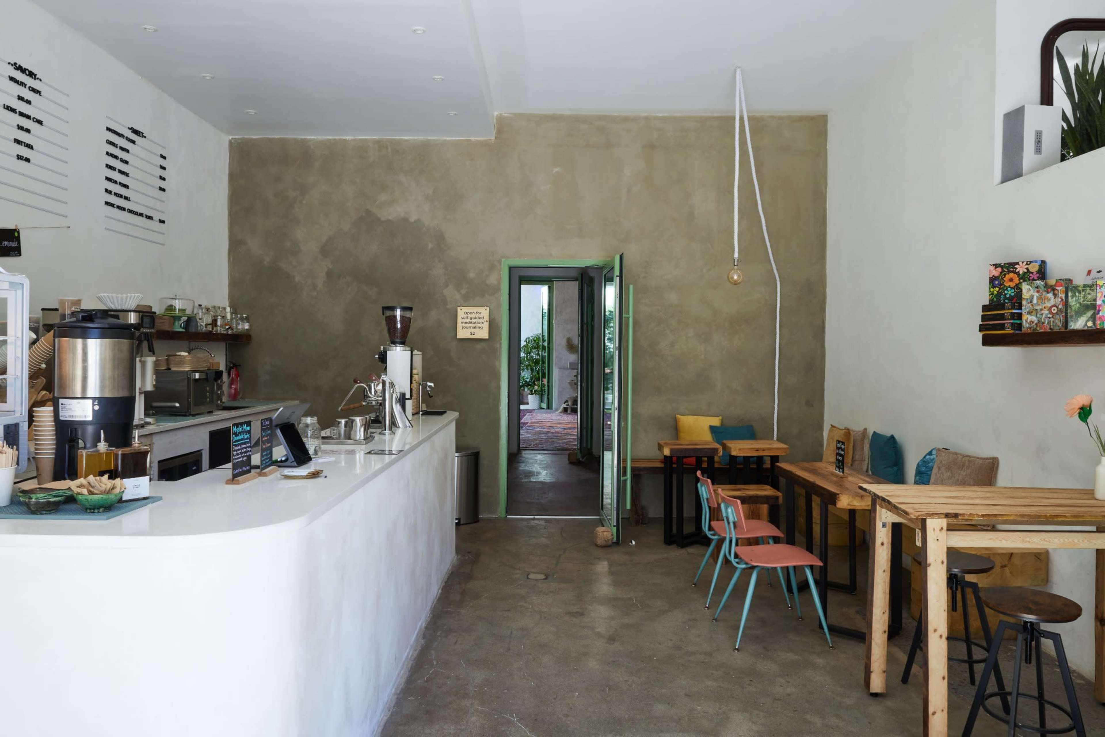 The image shows a minimalist café with a white counter, wooden tables, colorful chairs, and an open doorway leading to a green space.