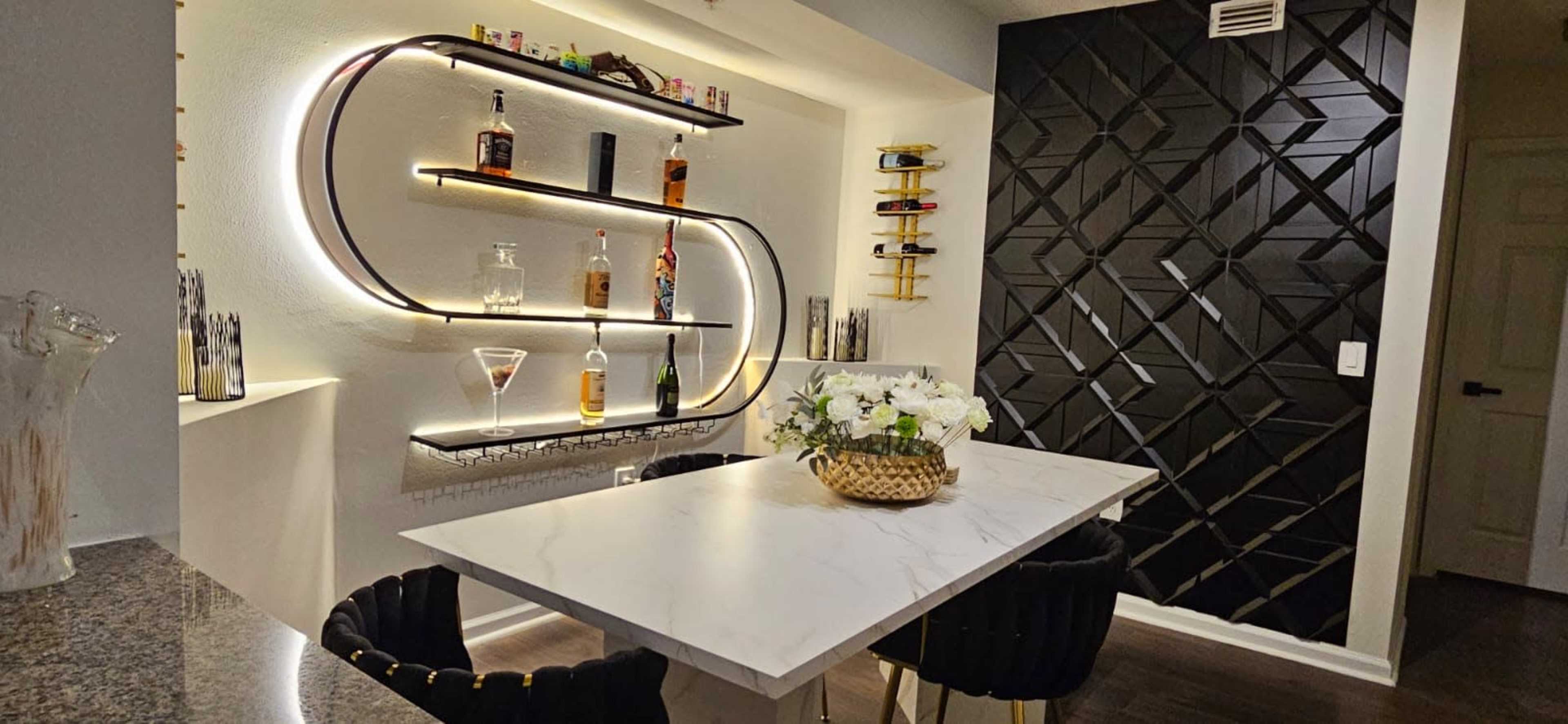 The image shows a contemporary bar area featuring illuminated shelves displaying liquor bottles and a marble-top table with black chairs, adjacent to a textured black wall.