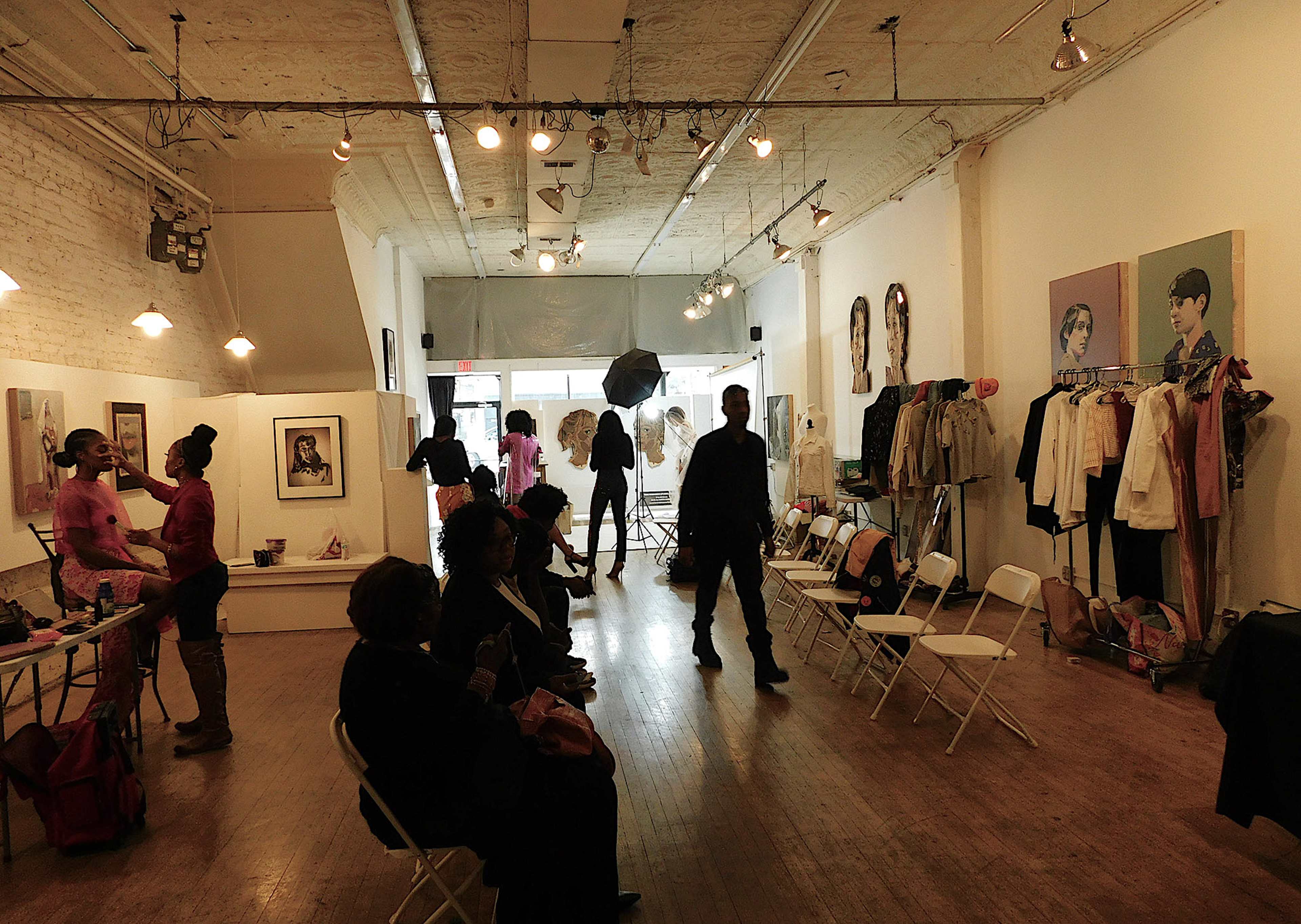 The image shows a modern art gallery with several people, chairs, and artworks displayed on the walls.