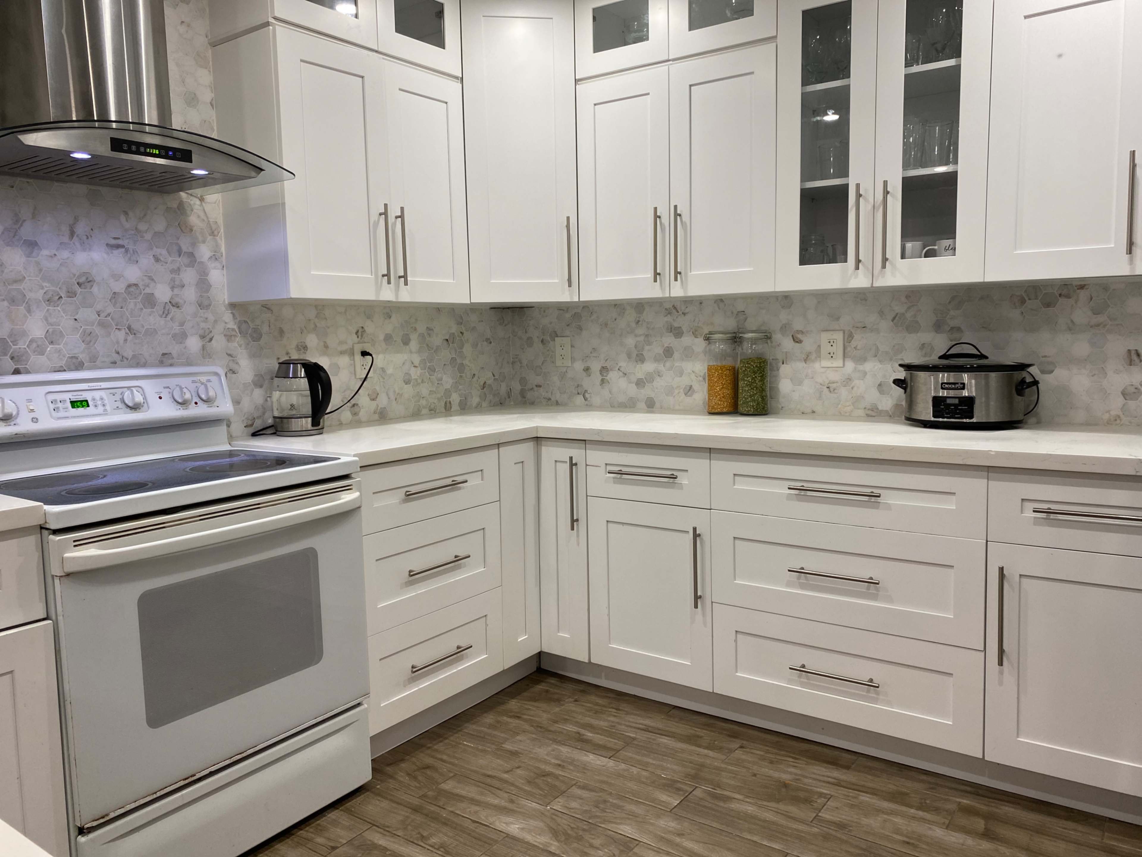 The image shows a modern kitchen with white cabinetry, a stove, and a counter featuring a kettle and a slow cooker.
