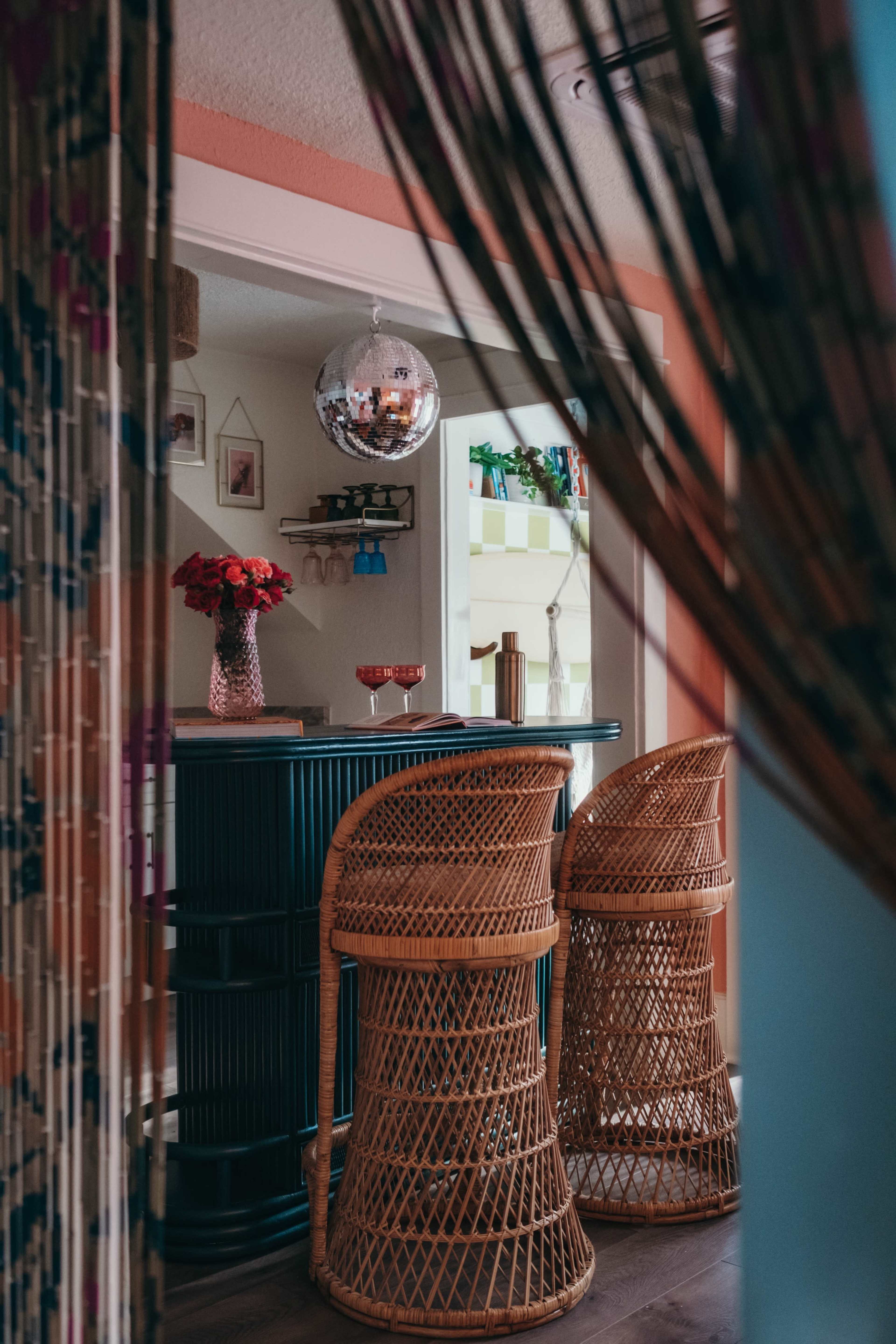 The image features a cozy bar area with two wicker stools, a disco ball hanging above, and flower arrangements on the countertop.