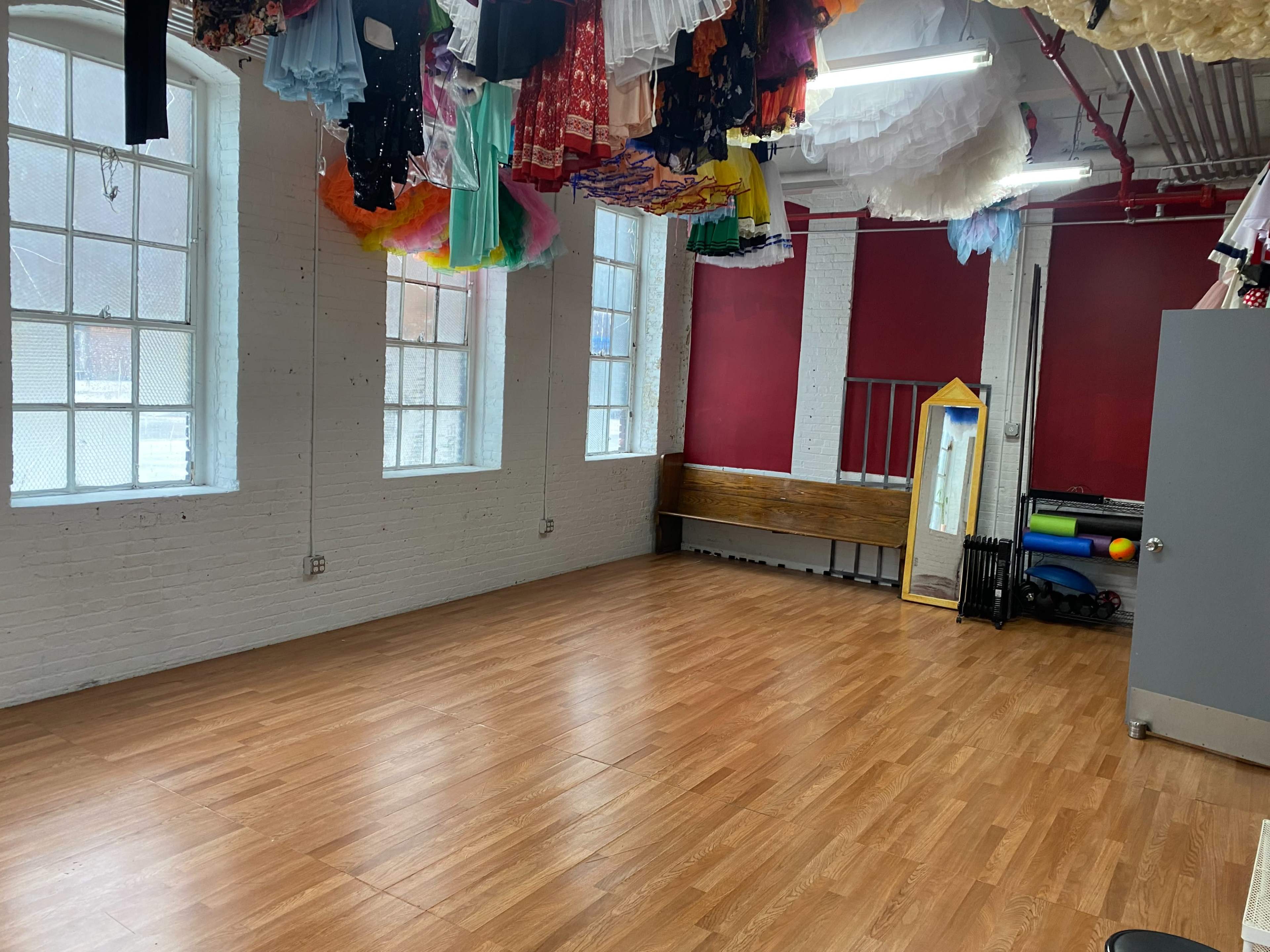 The room features wooden flooring, large windows, and a variety of costumes hanging from the ceiling, with a mirror and exercise equipment against one wall.