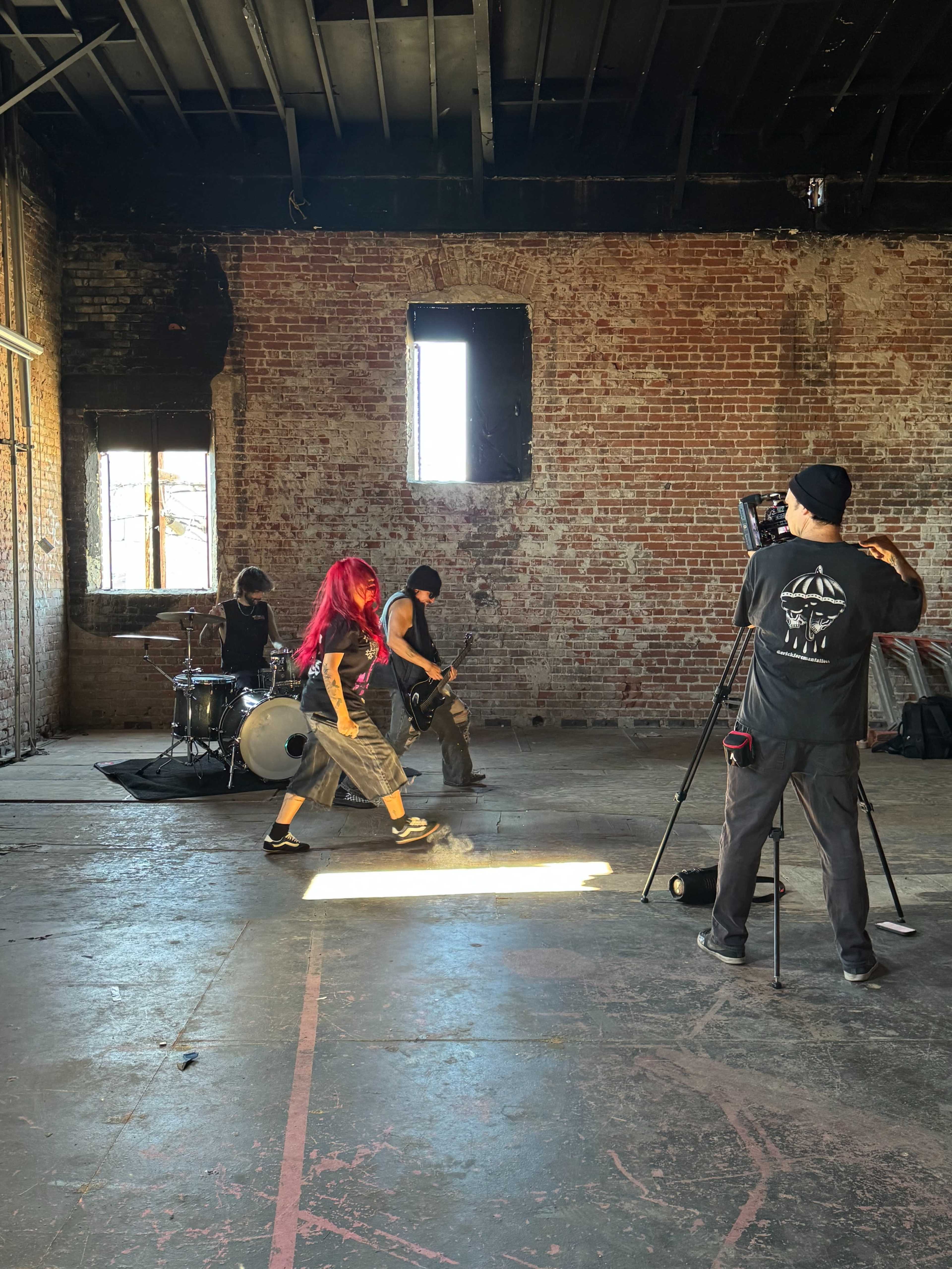 A band performs in a brick-walled room with a camera operator capturing the scene.
