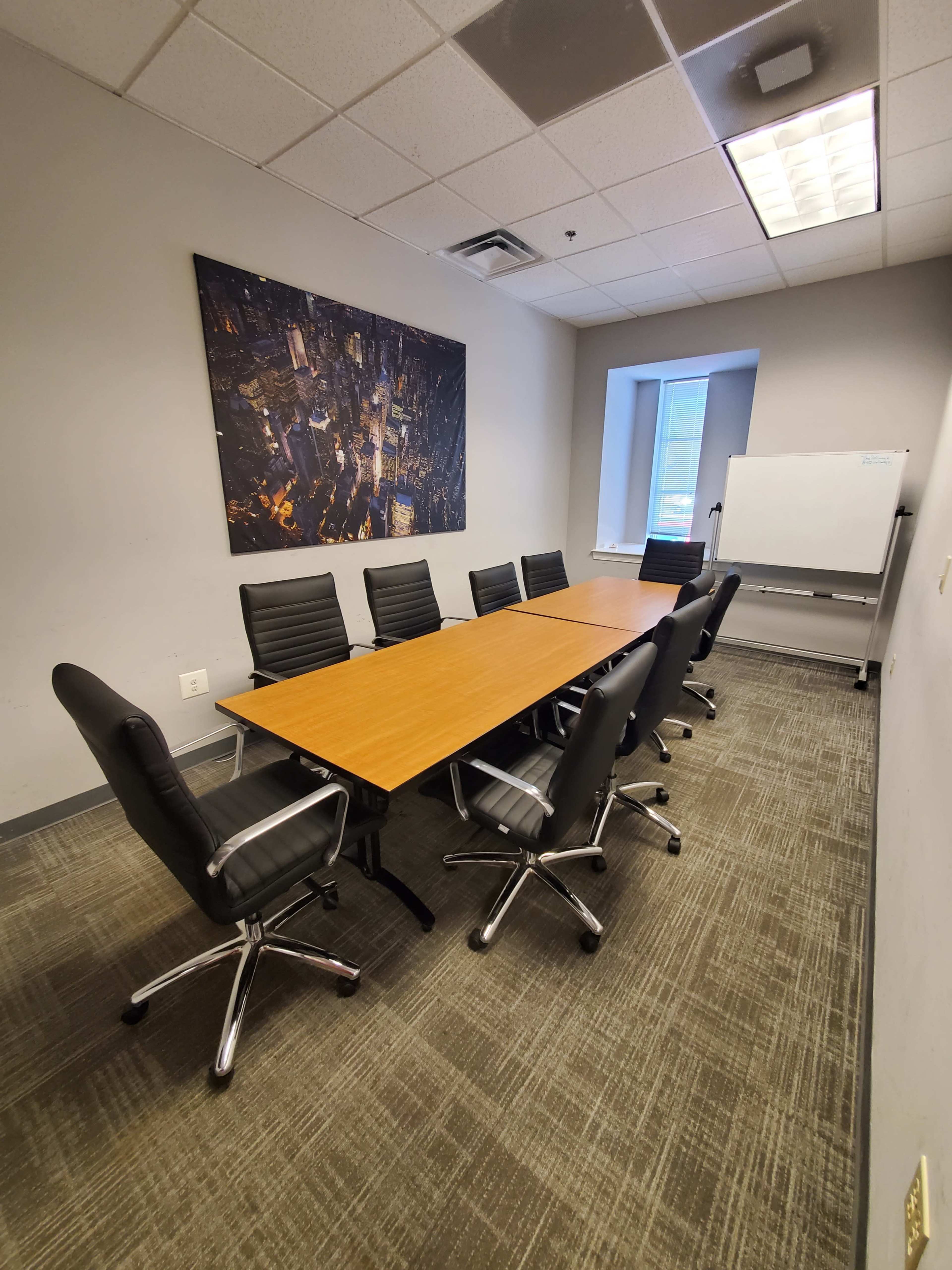 A conference room with a long wooden table surrounded by eight black office chairs, and a large artwork on the wall, along with a whiteboard and a window.