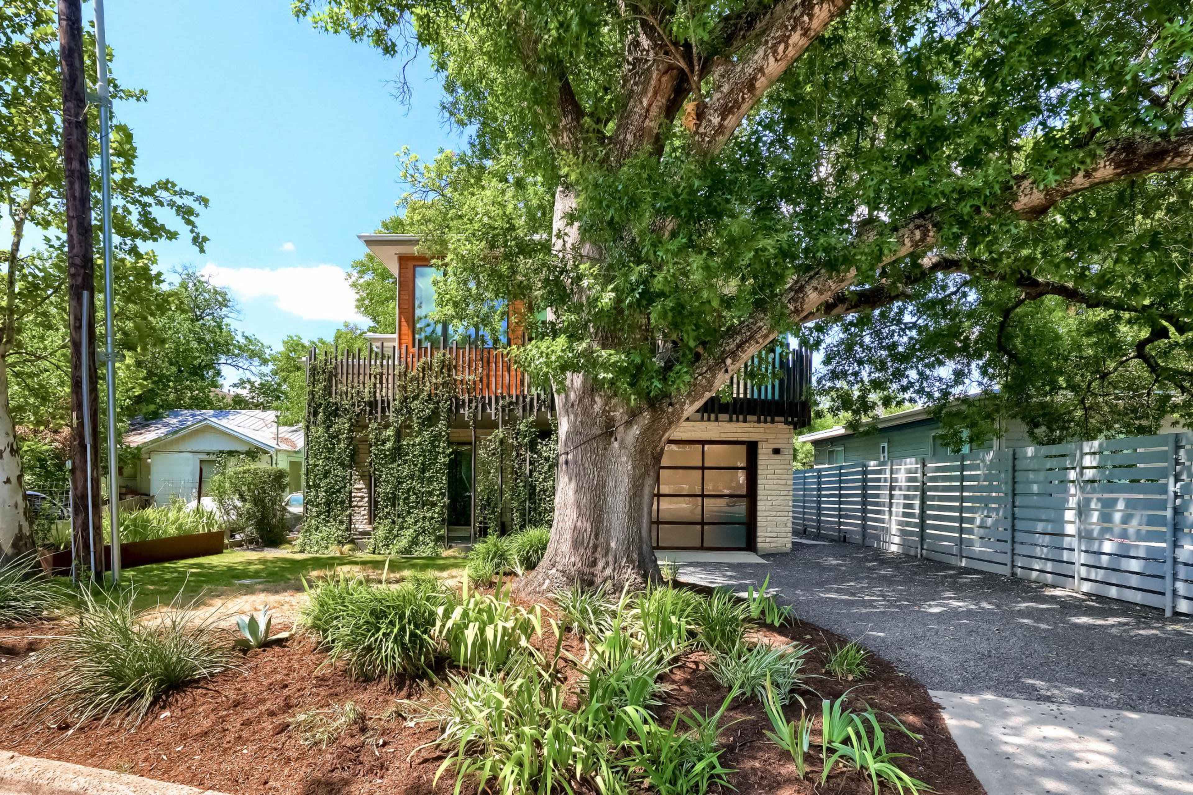 A modern two-story house is shaded by a large tree, surrounded by landscaped greenery and gravel pathways.