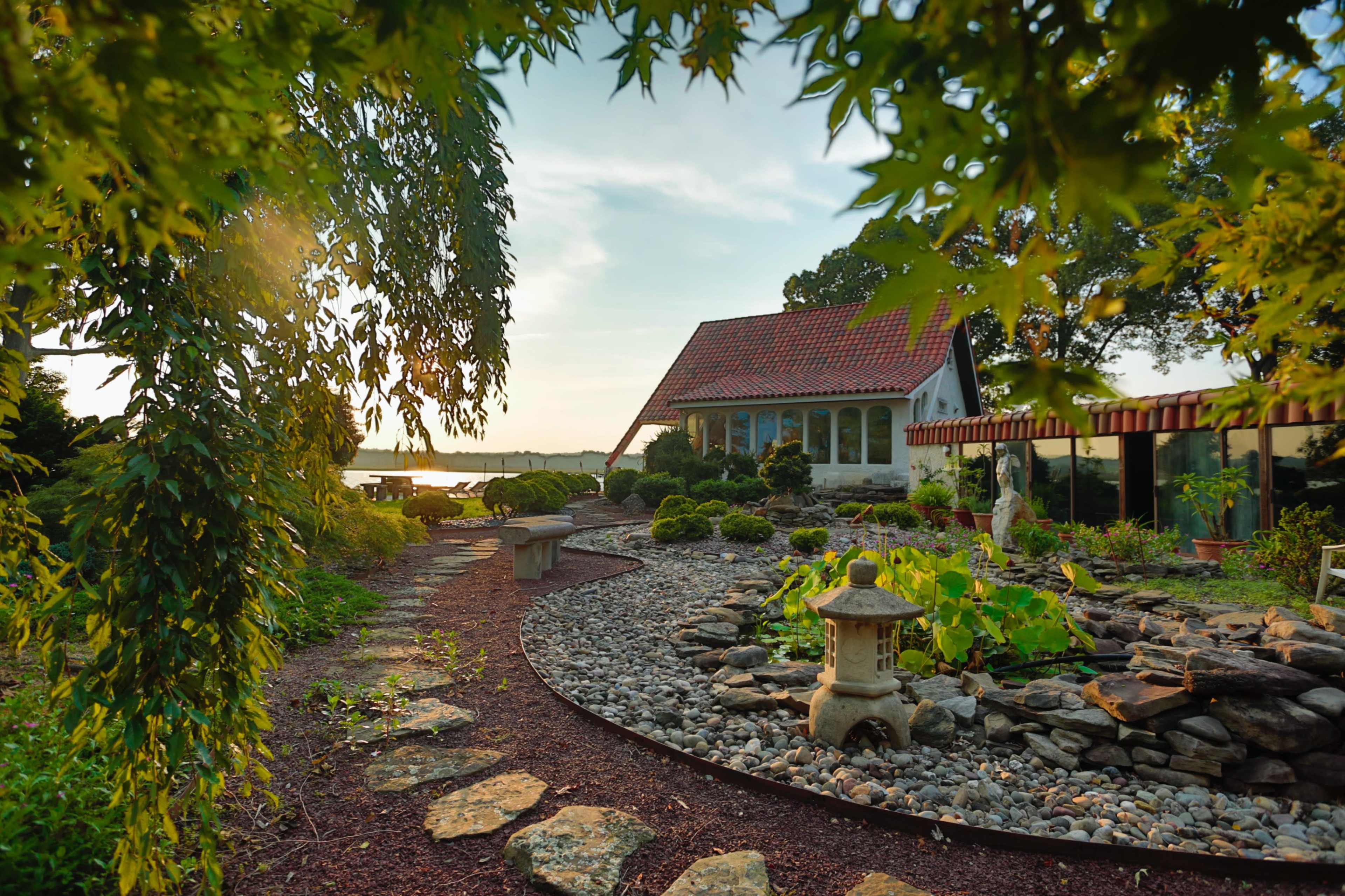 A garden path winds through a landscaped area with a stone lantern and a view of a house near a body of water.