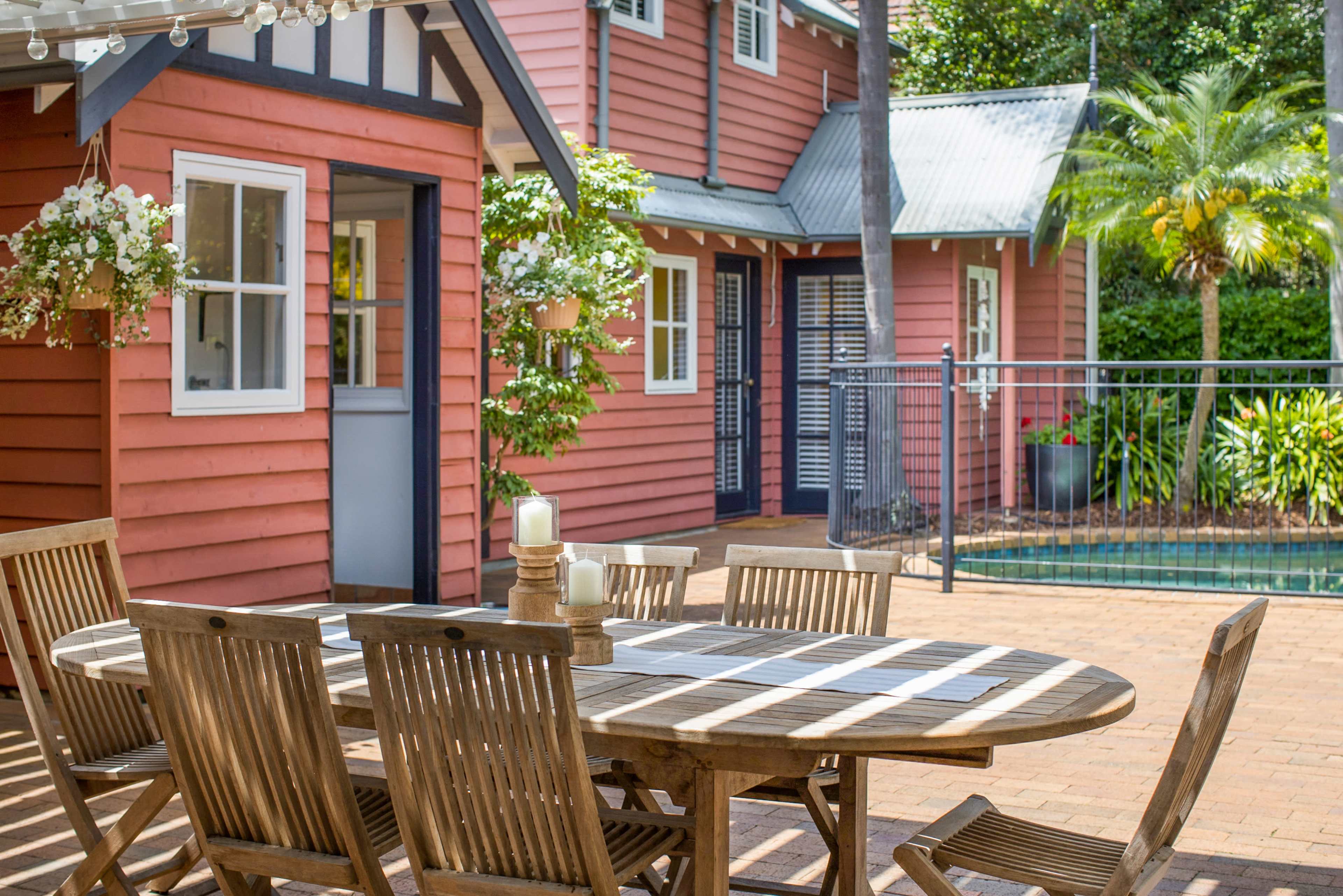 A wooden dining table with chairs is set up on a patio next to a pink house and a swimming pool surrounded by greenery.