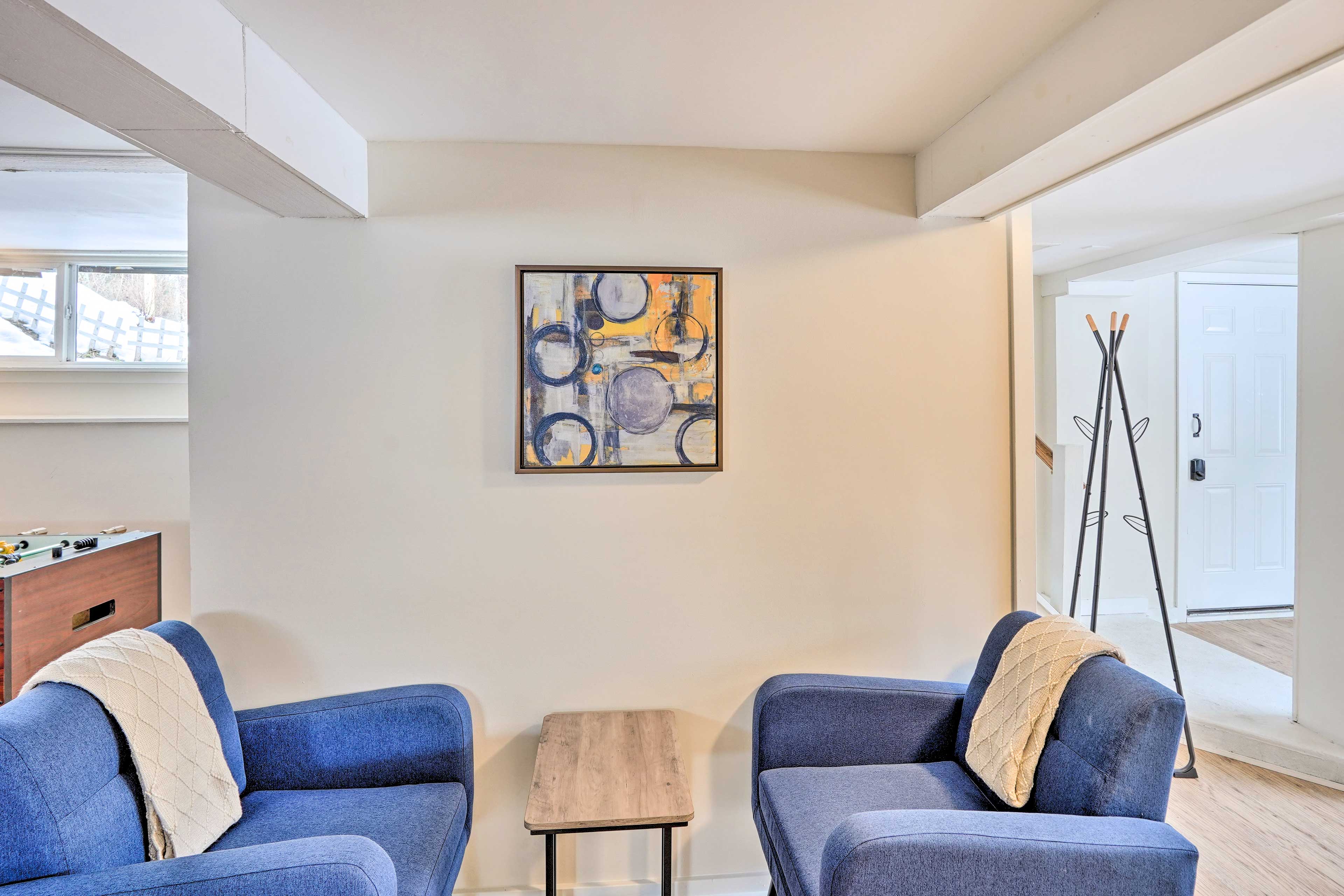A pair of blue armchairs with cream-colored throw blankets sit on either side of a small wooden table in a room featuring a piece of abstract wall art.