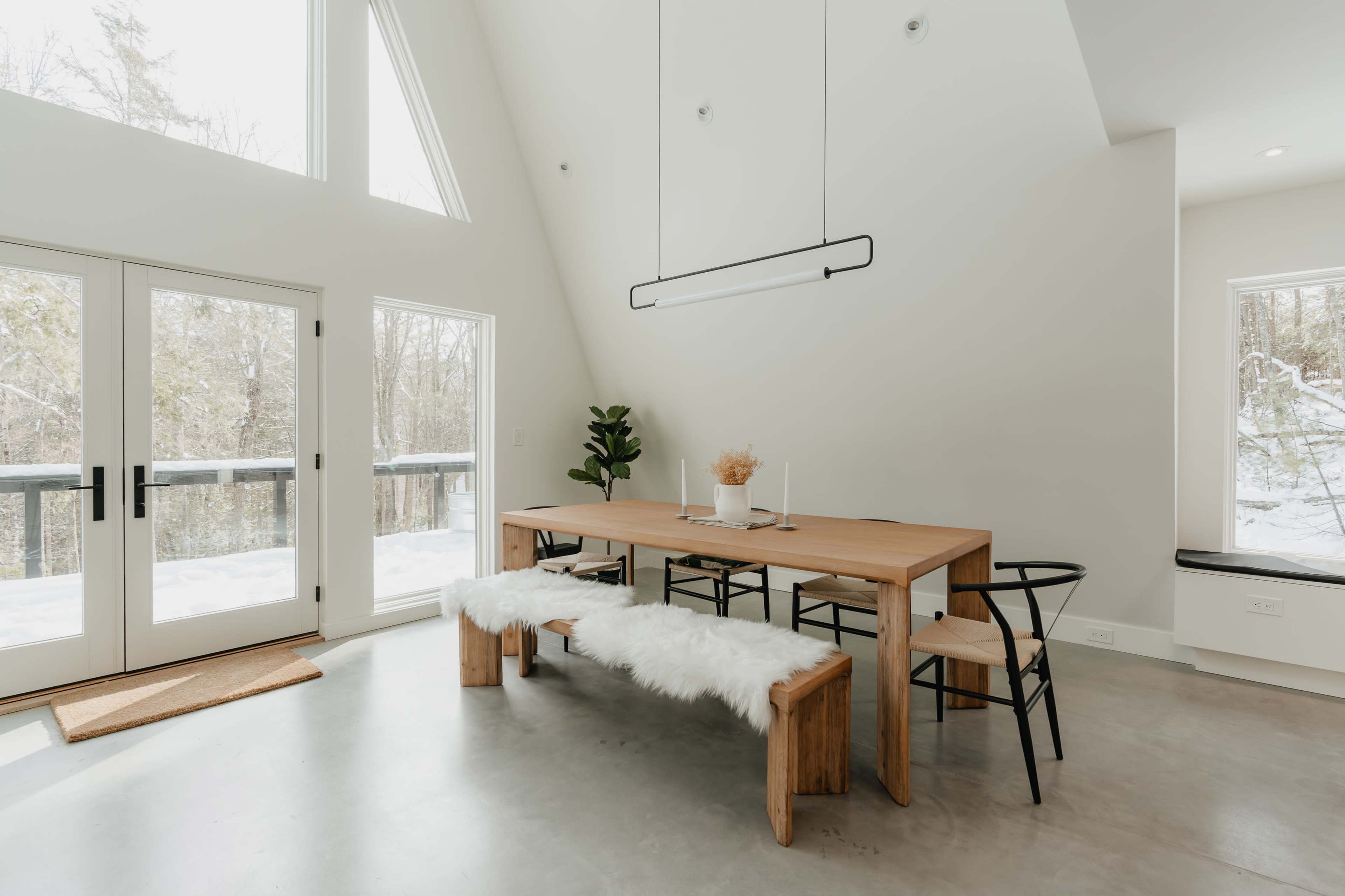 A spacious dining area features a wooden table with benches and black chairs, surrounded by large windows that provide natural light and views of a snowy forest.