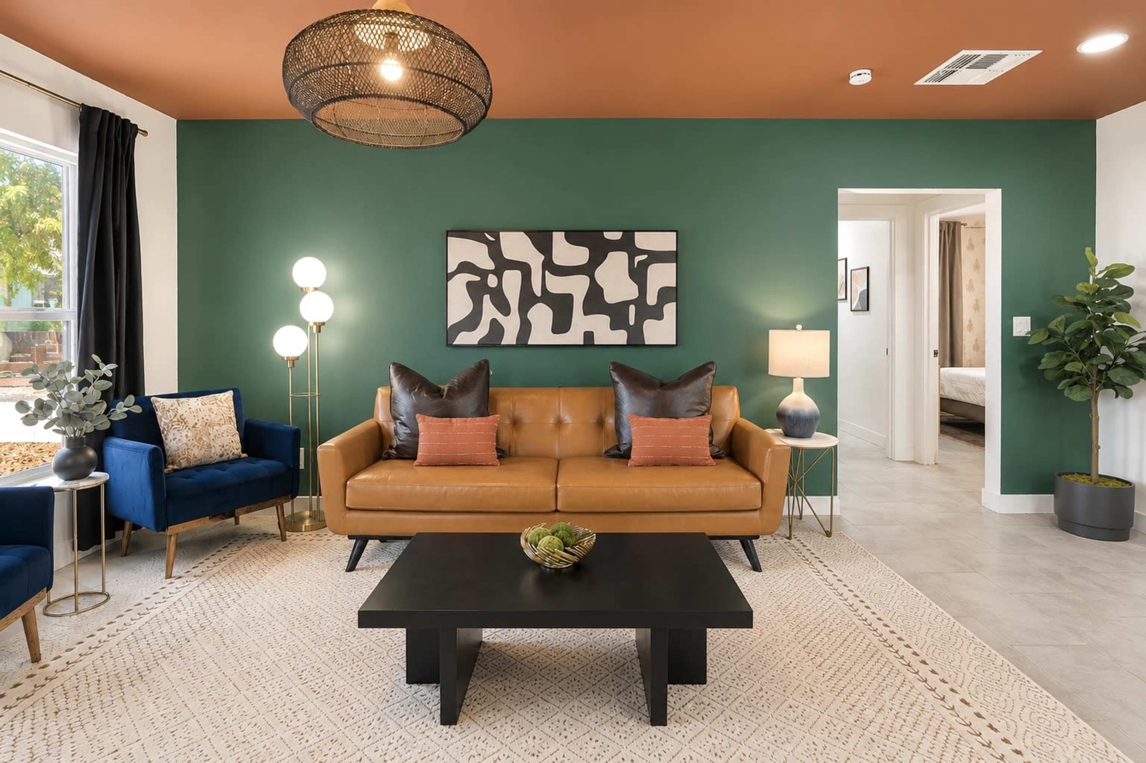 A modern living room features a brown leather sofa with orange and black pillows, surrounded by blue chairs and a black coffee table, against a green wall with an abstract artwork.
